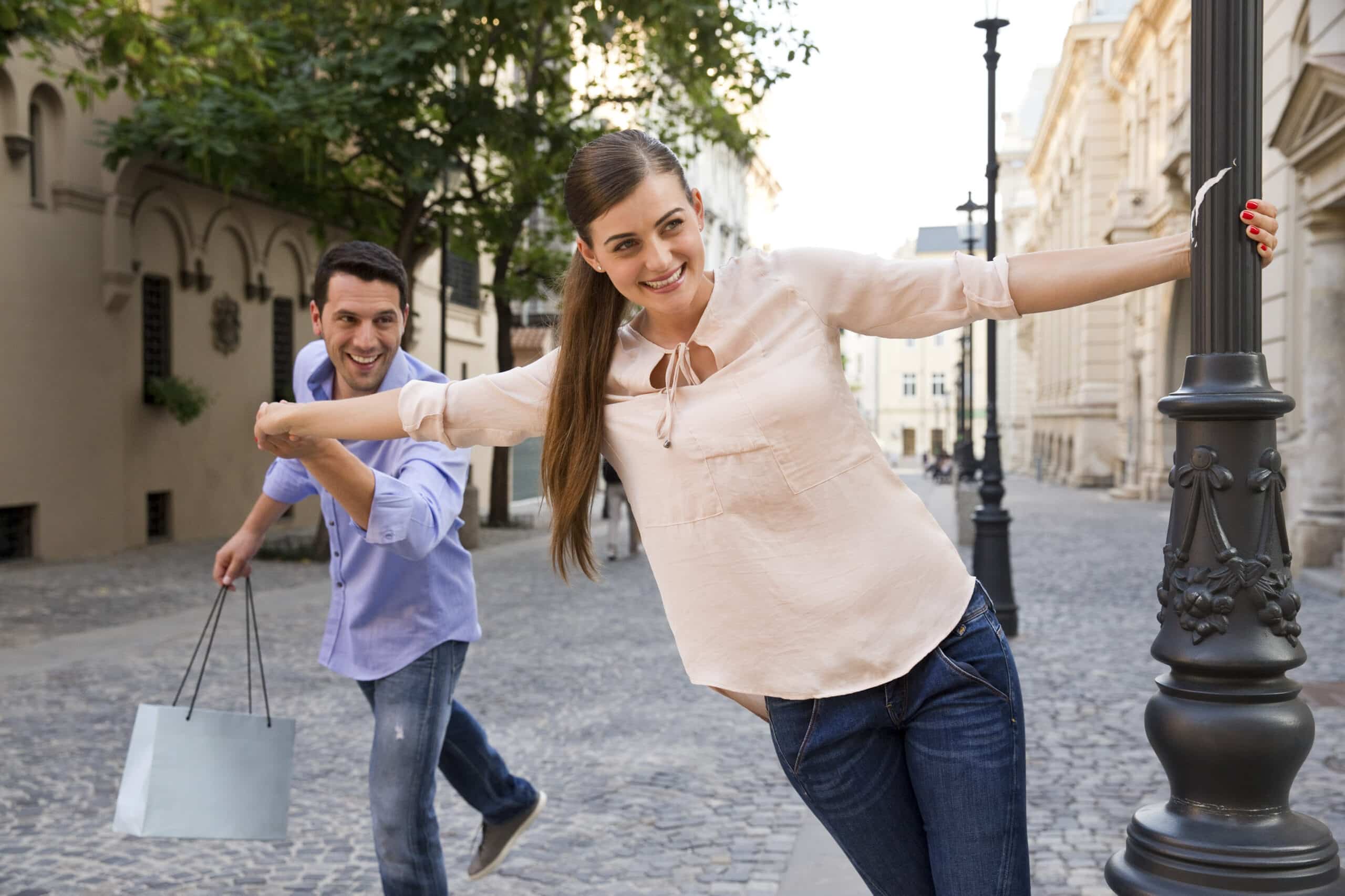 Eine fröhliche Frau schwingt sich um einen Laternenpfahl auf einer Stadtstraße und lächelt in die Kamera, während ein Mann ihr spielerisch mit einer Einkaufstüte in der Hand nachjagt. Warmes Sonnenlicht erhellt die Szene. © Fotografie Tomas Rodriguez