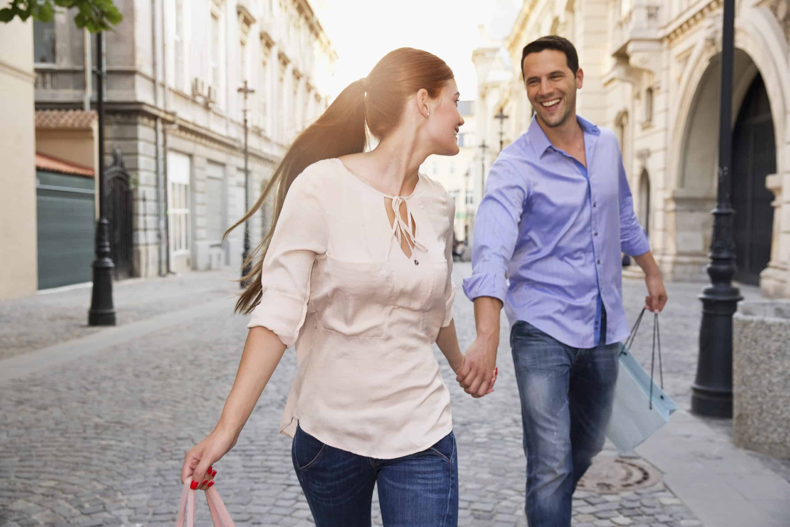 Ein fröhlicher Mann und eine fröhliche Frau gehen Händchen haltend durch eine sonnige Stadtstraße, tragen Einkaufstüten und schauen sich lächelnd an. © Fotografie Tomas Rodriguez