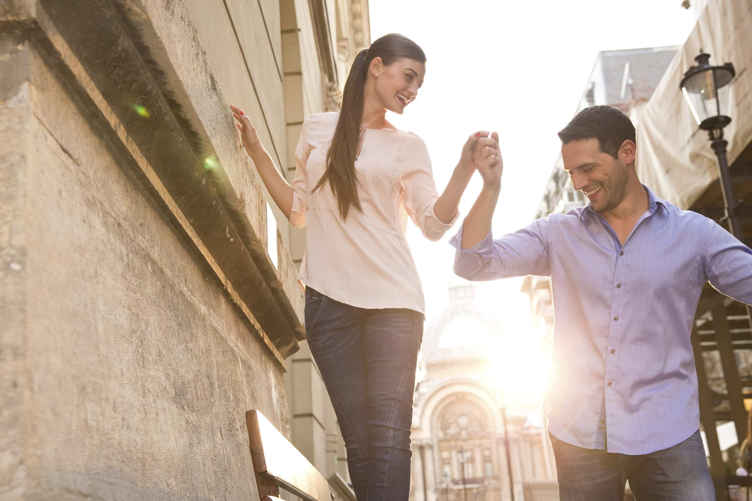 Ein fröhliches Paar tanzt auf einer sonnenbeschienenen Stadtstraße. Die Frau führt den Mann an der Hand und hält ihn. Beide lächeln breit vor der malerischen städtischen Kulisse. © Fotografie Tomas Rodriguez