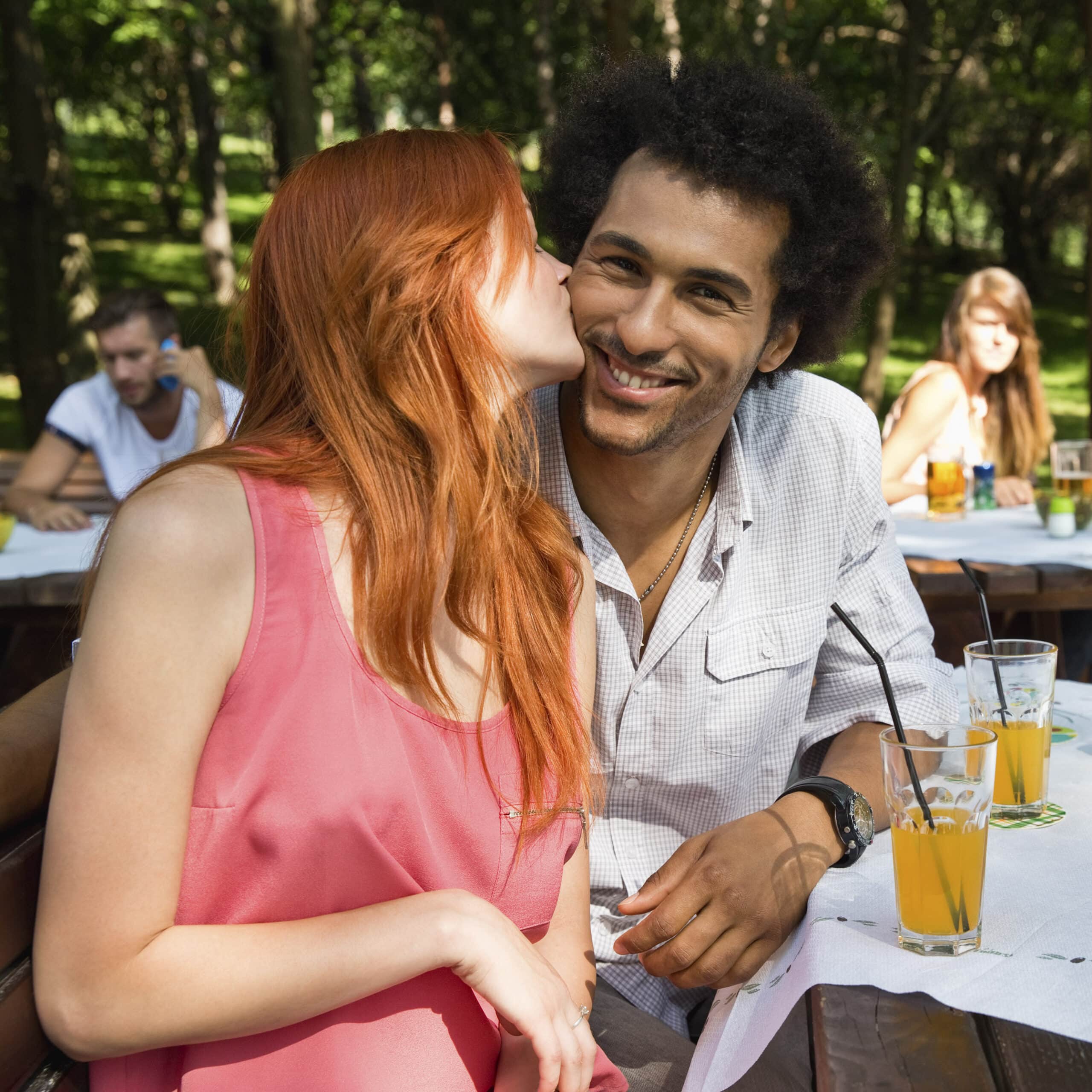 Eine Frau mit roten Haaren küsst einen lächelnden Mann mit lockigem Haar an einem sonnigen Picknicktisch im Park auf die Wange, im Hintergrund sind andere Menschen zu sehen. Getränke und ein sonniger Tag tragen zu der freudigen Szene bei. © Fotografie Tomas Rodriguez