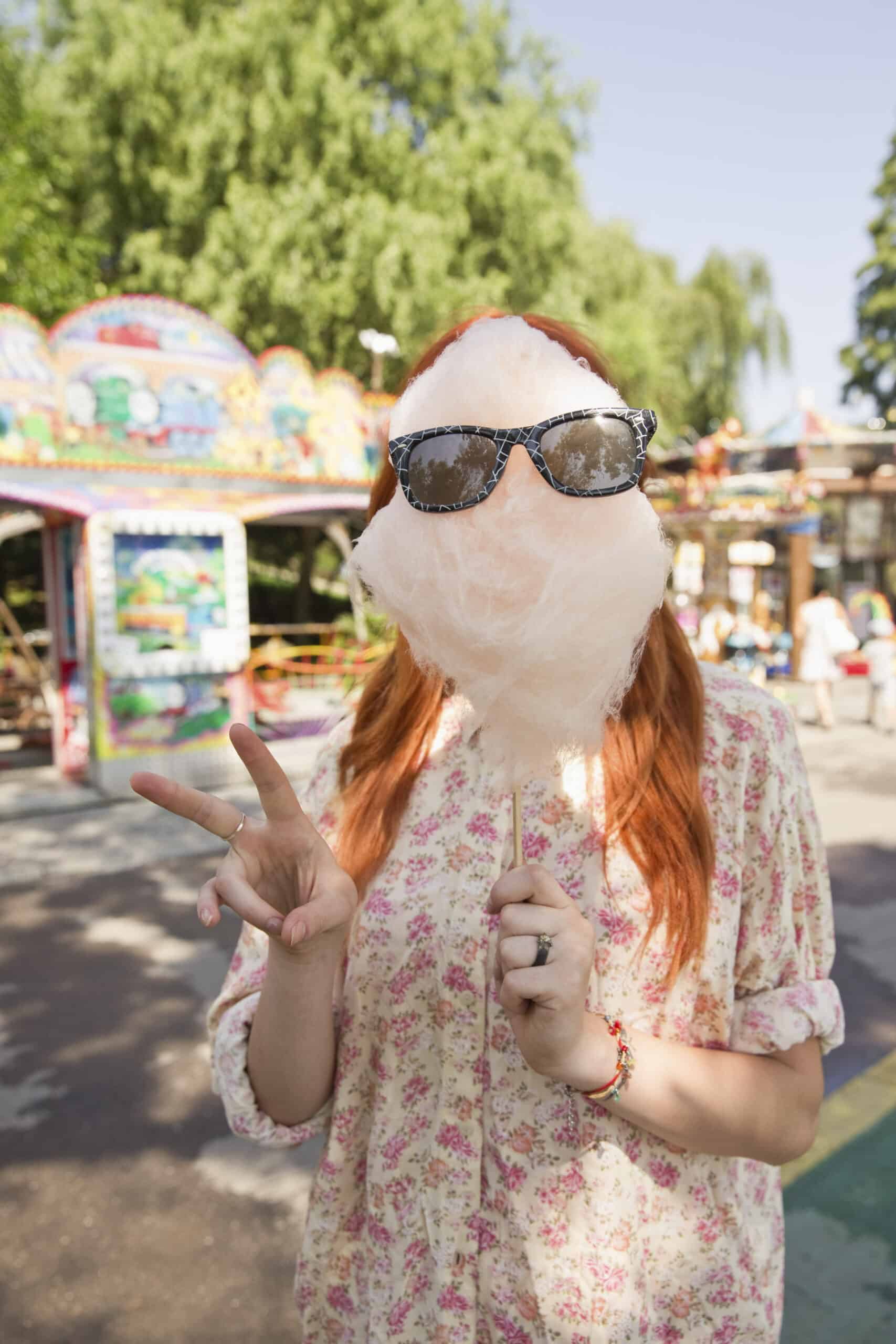 Eine Frau mit roten Haaren und einer Sonnenbrille, die teilweise von einer großen Zuckerwatte verdeckt wird, macht in einem Vergnügungspark ein Peace-Zeichen. © Fotografie Tomas Rodriguez