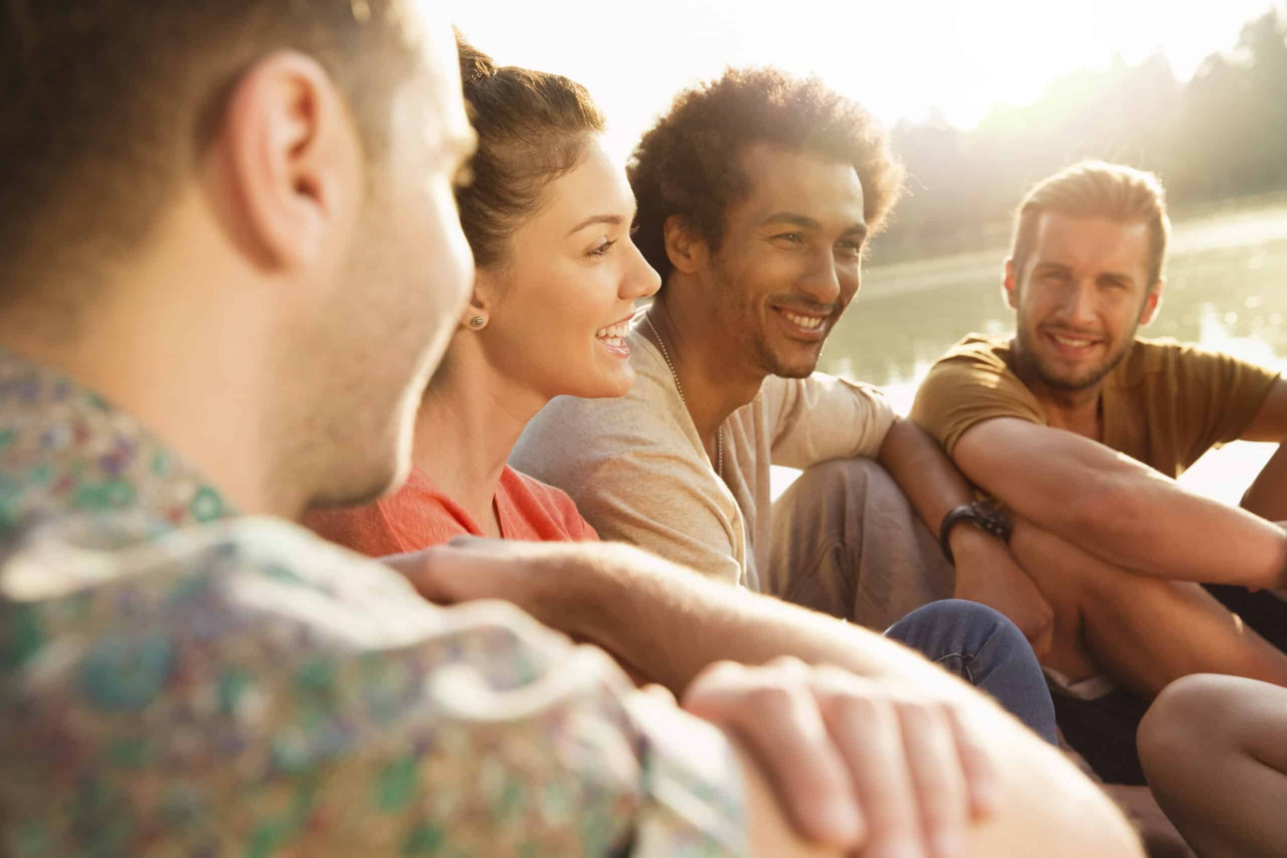 Vier Freunde genießen einen sonnigen Tag im Freien. Sie sitzen eng beieinander, lächeln und unterhalten sich fröhlich. Das Sonnenlicht fällt sanft auf ihre Gesichter und unterstreicht ihre freudigen Ausdrücke. © Fotografie Tomas Rodriguez