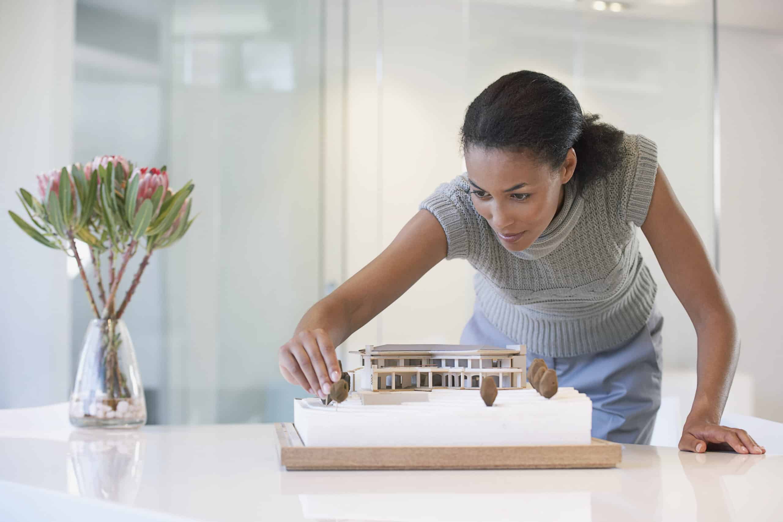 Eine Afroamerikanerin in einem grauen ärmellosen Top lehnt sich über einen Schreibtisch, um ein detailliertes Architekturmodell in einem hellen, modernen Büro zu untersuchen. © Fotografie Tomas Rodriguez