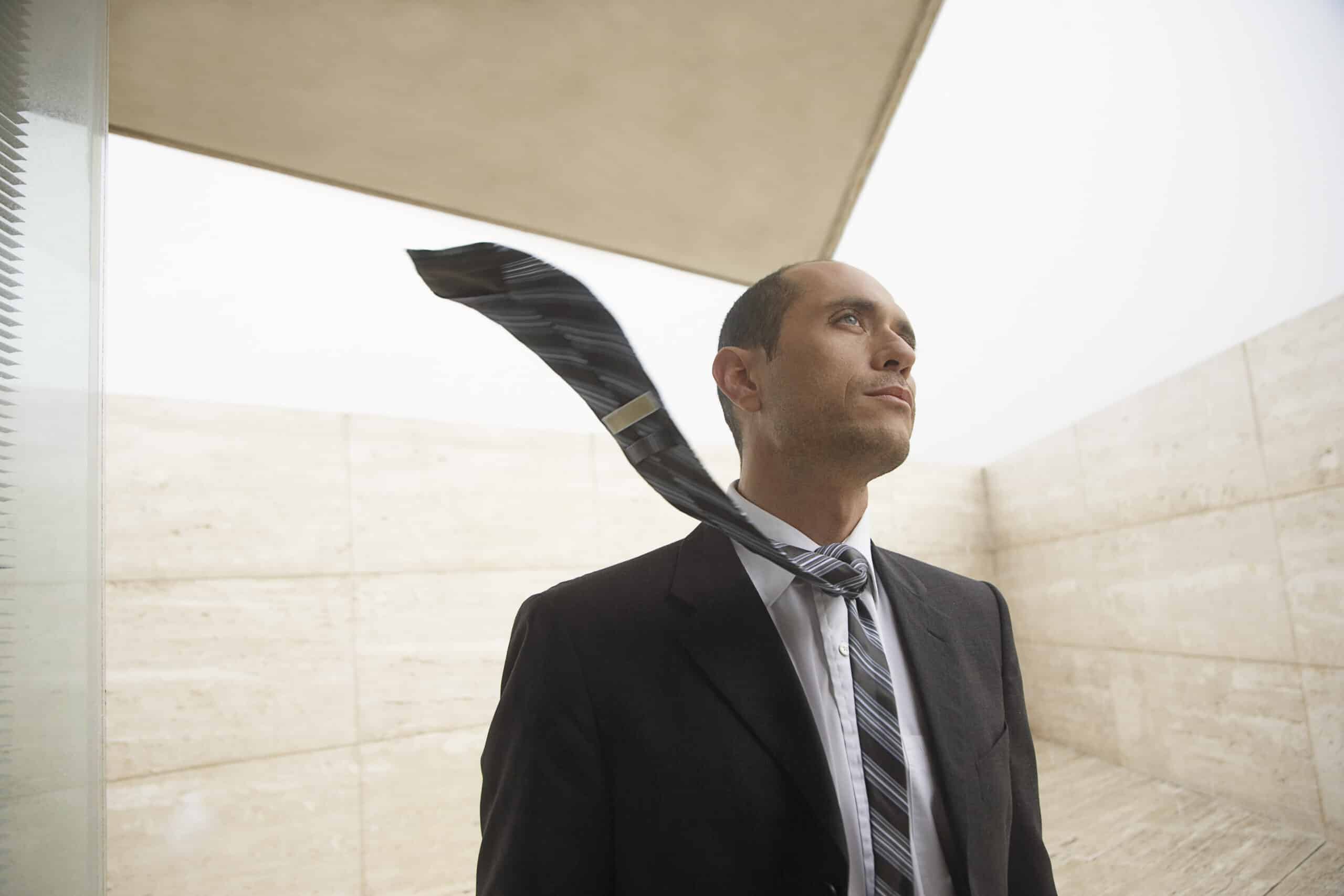 Ein Mann im Businessanzug, dessen Krawatte im Wind flattert, steht vor einem minimalistischen Betonhintergrund und blickt nachdenklich nach oben. © Fotografie Tomas Rodriguez