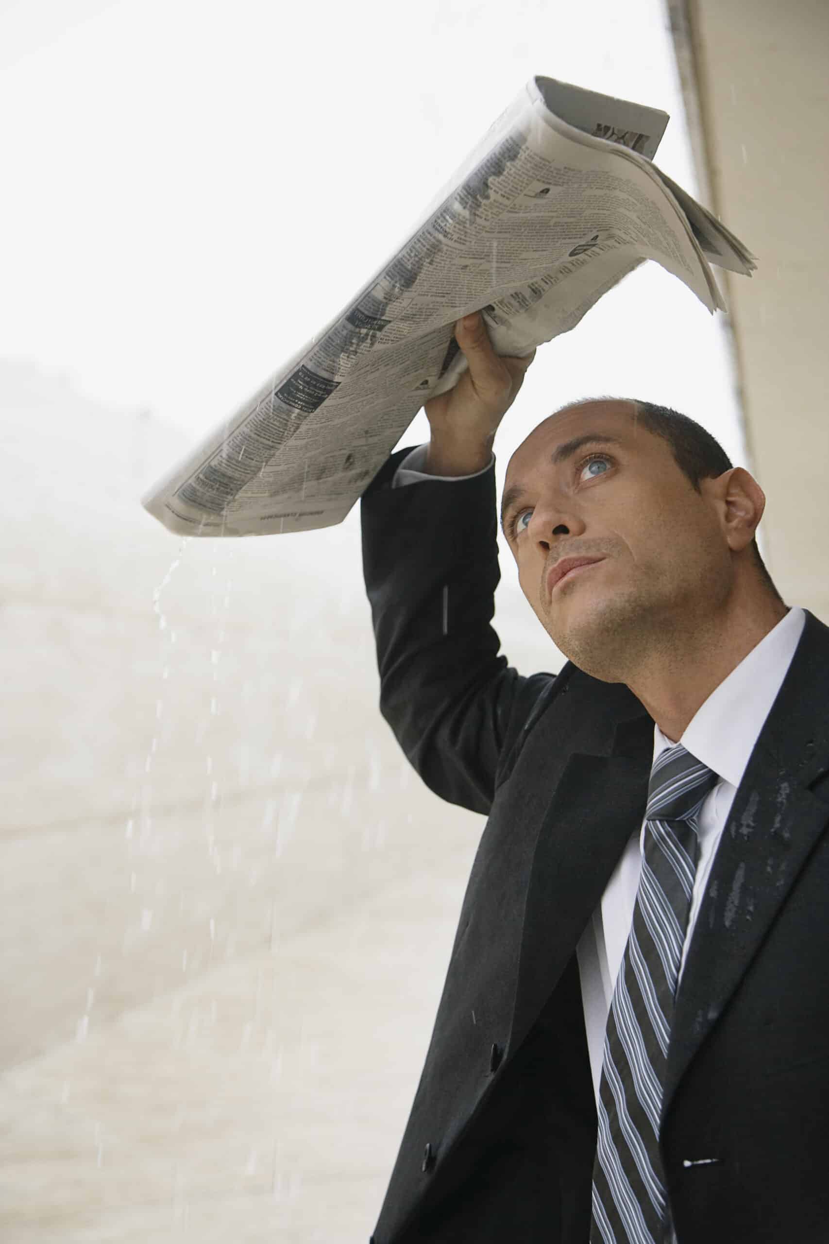 Ein Mann im Anzug benutzt eine Zeitung als improvisierten Regenschirm im Regen und blickt mit besorgtem Gesichtsausdruck nach oben. © Fotografie Tomas Rodriguez