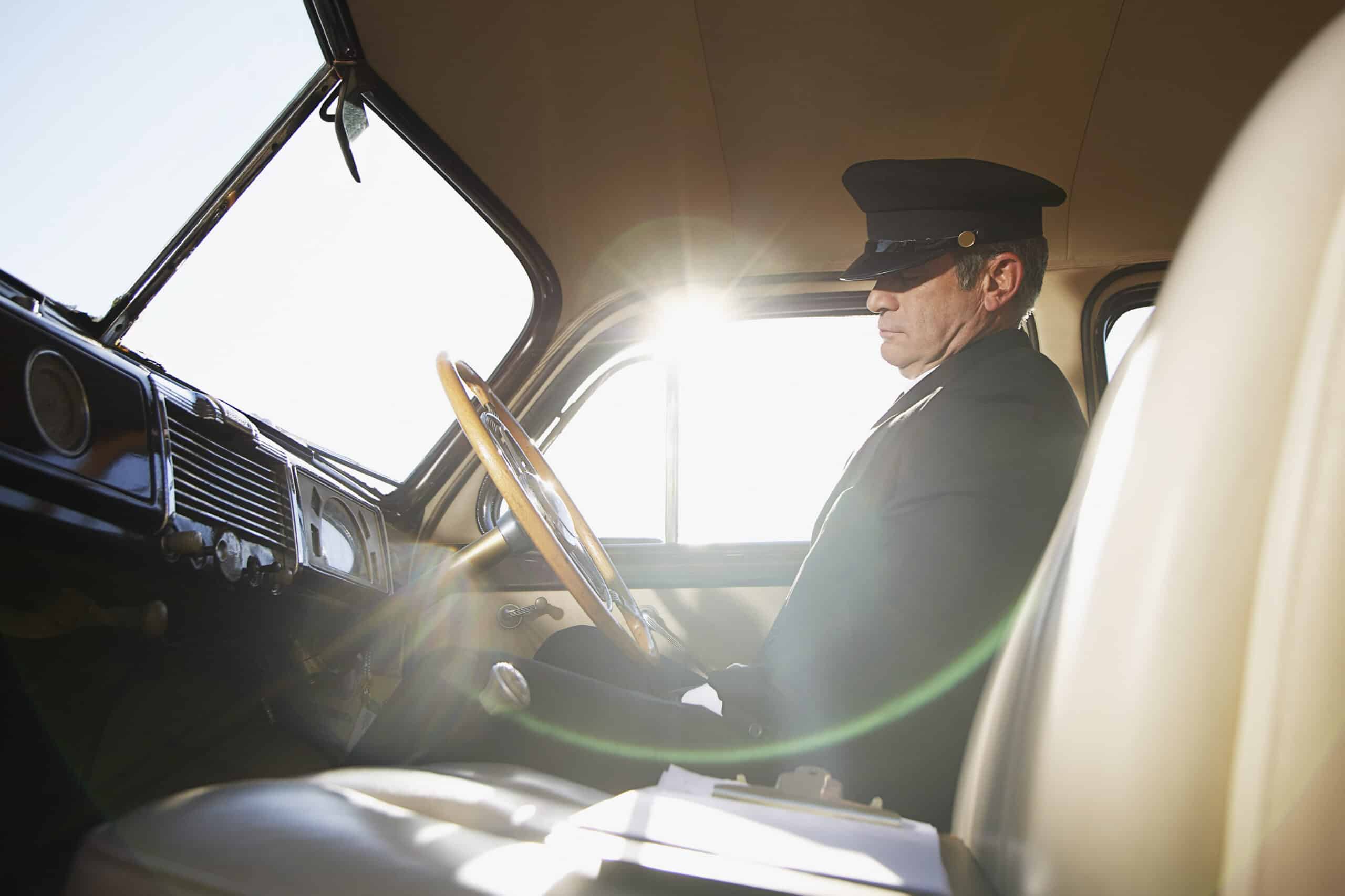 Ein Chauffeur in Uniform und Sonnenbrille fährt einen Oldtimer, der in Sonnenlicht getaucht ist und seinen Blick fest auf die Straße vor ihm gerichtet hat. © Fotografie Tomas Rodriguez