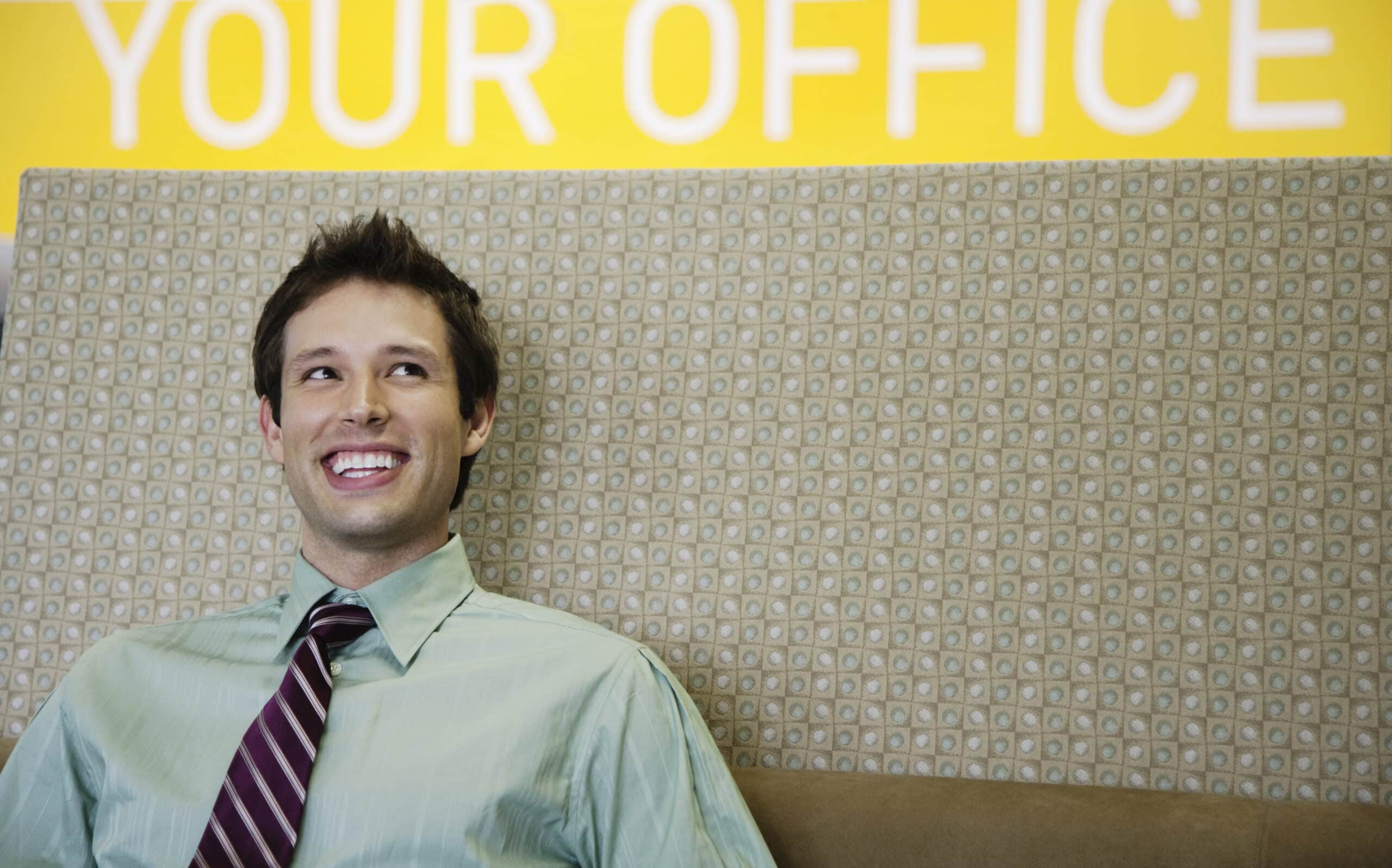 Ein fröhlicher Mann in Hemd und Krawatte sitzt vor einer Wand mit einem Schild mit der Aufschrift „Ihr Büro“, lächelt und blickt nach links oben. © Fotografie Tomas Rodriguez