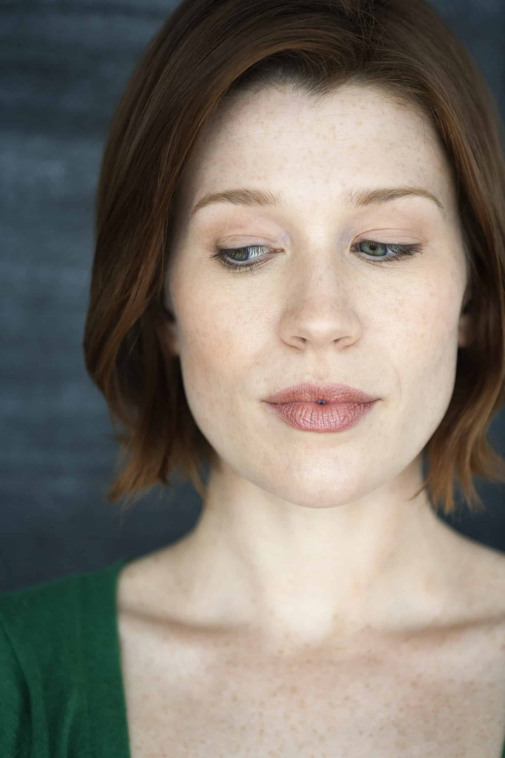 Porträt einer Frau mit kurzem rotbraunem Haar, hellen Sommersprossen und dezentem Make-up. Sie trägt ein grünes Oberteil und blickt mit sanftem Gesichtsausdruck vor einem grauen Hintergrund nach unten. © Fotografie Tomas Rodriguez