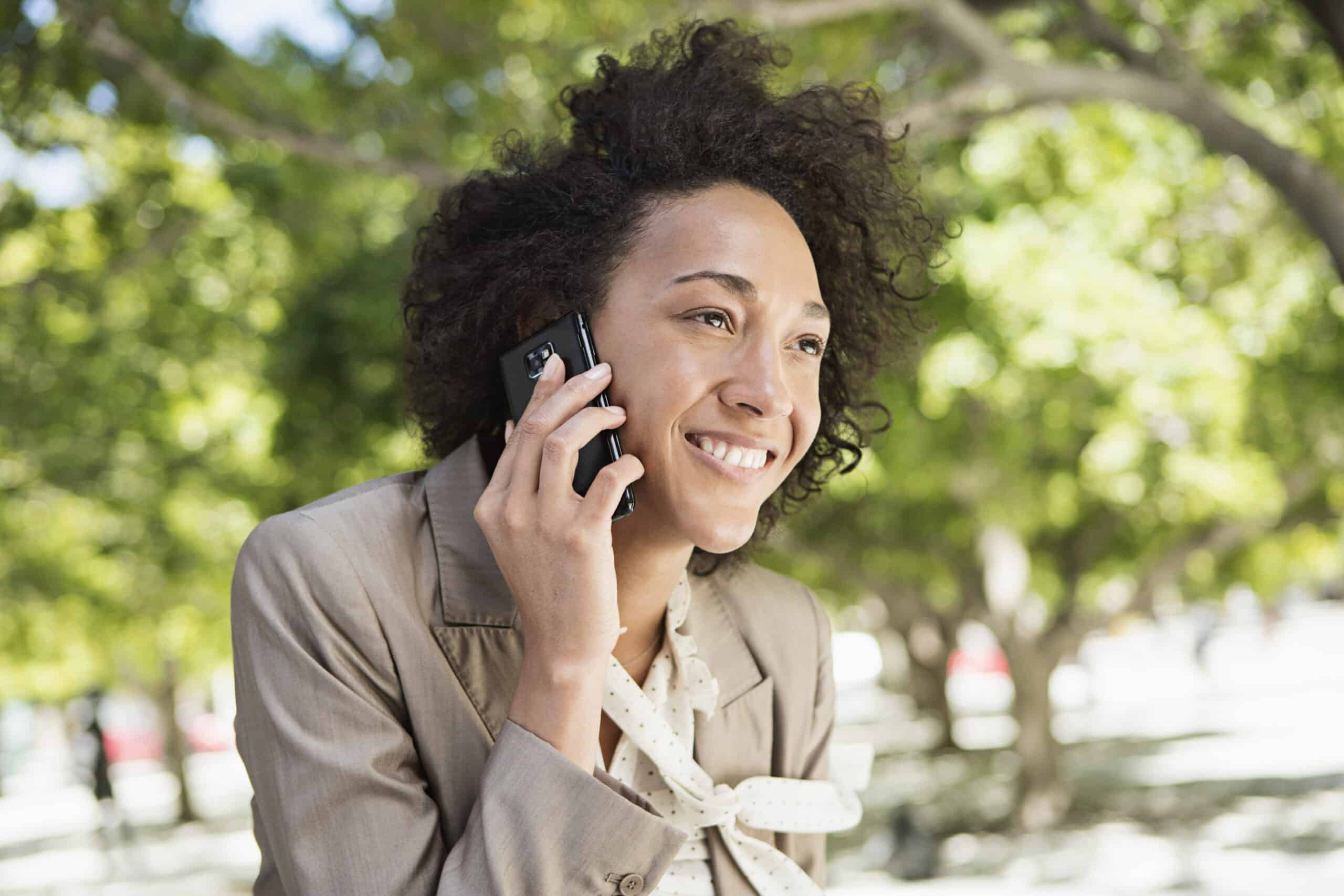 Eine fröhliche Frau mit lockigem Haar telefoniert mit einem Smartphone in einem sonnigen Park, umgeben von grünen Bäumen. Sie trägt eine hellbraune Jacke und eine weiße Bluse. © Fotografie Tomas Rodriguez