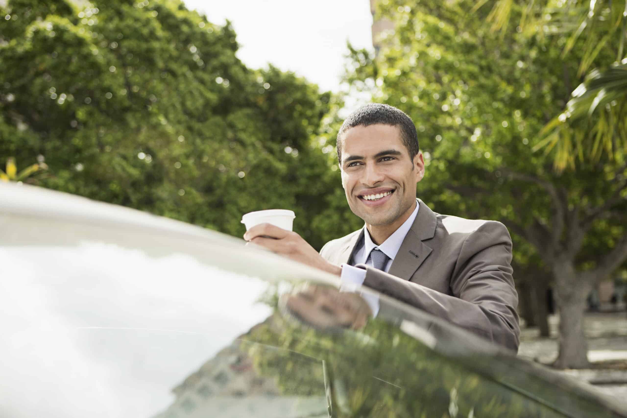 Ein lächelnder Geschäftsmann mit einer Kaffeetasse in der Hand lehnt in einer sonnigen Parklandschaft, umgeben von üppigem Grün, an einem Auto. © Fotografie Tomas Rodriguez