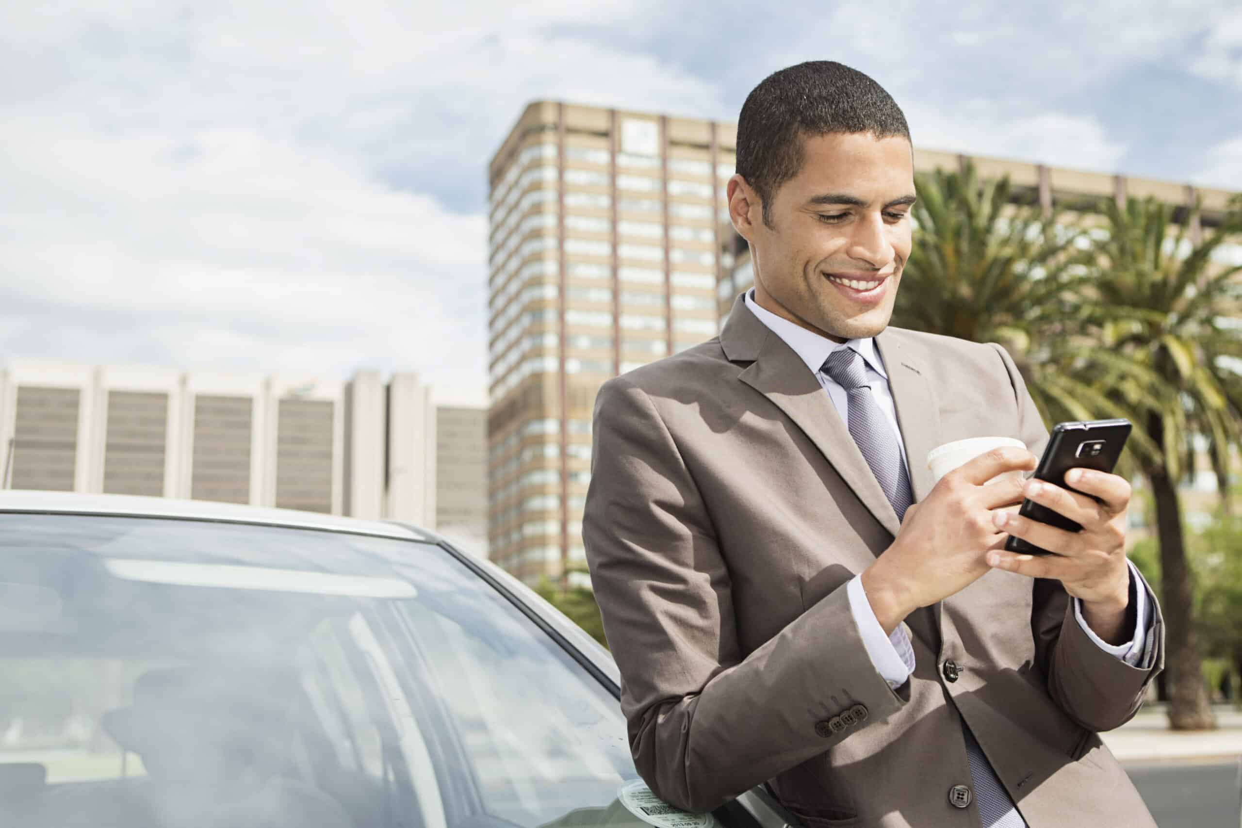 Ein lächelnder Geschäftsmann im grauen Anzug steht neben einem Auto und benutzt sein Smartphone mit einer modernen Stadtlandschaft im Hintergrund. © Fotografie Tomas Rodriguez