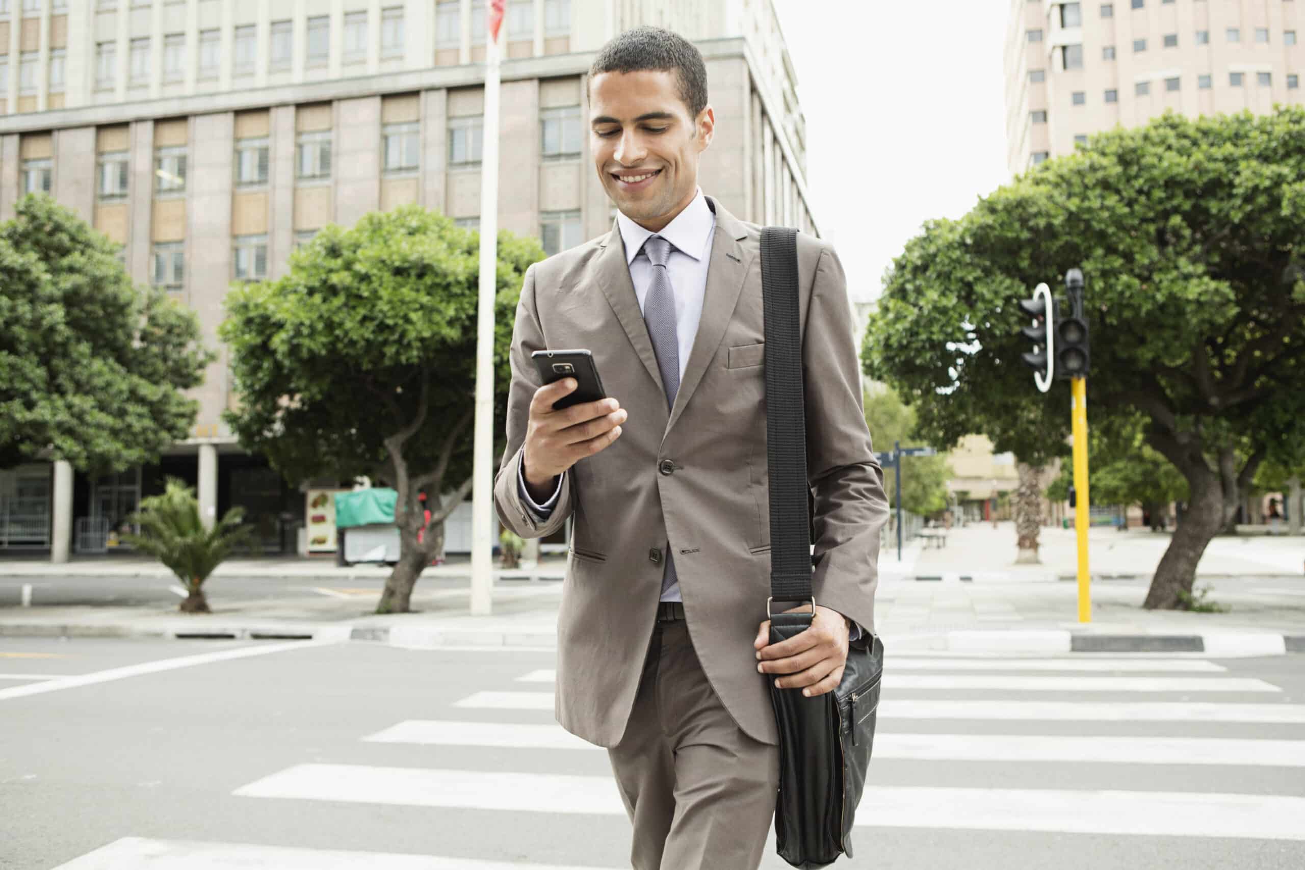 Ein lächelnder junger Geschäftsmann im Anzug, der ein Smartphone und eine Umhängetasche in der Hand hält, geht über eine Stadtstraße mit Bäumen und Gebäuden im Hintergrund. © Fotografie Tomas Rodriguez