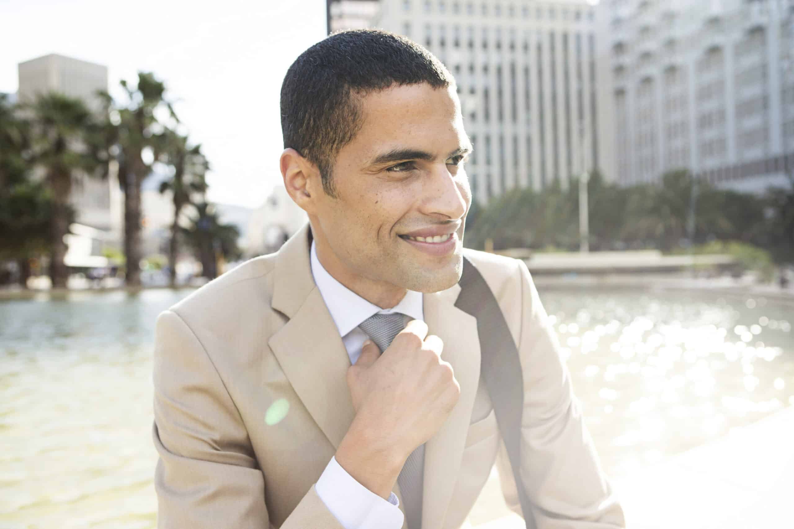 Ein lächelnder Geschäftsmann im beigen Anzug richtet seine Krawatte. Er steht im Freien mit einer Stadtlandschaft und einem Brunnen im Hintergrund und strahlt ein selbstbewusstes und entspanntes Auftreten aus. © Fotografie Tomas Rodriguez
