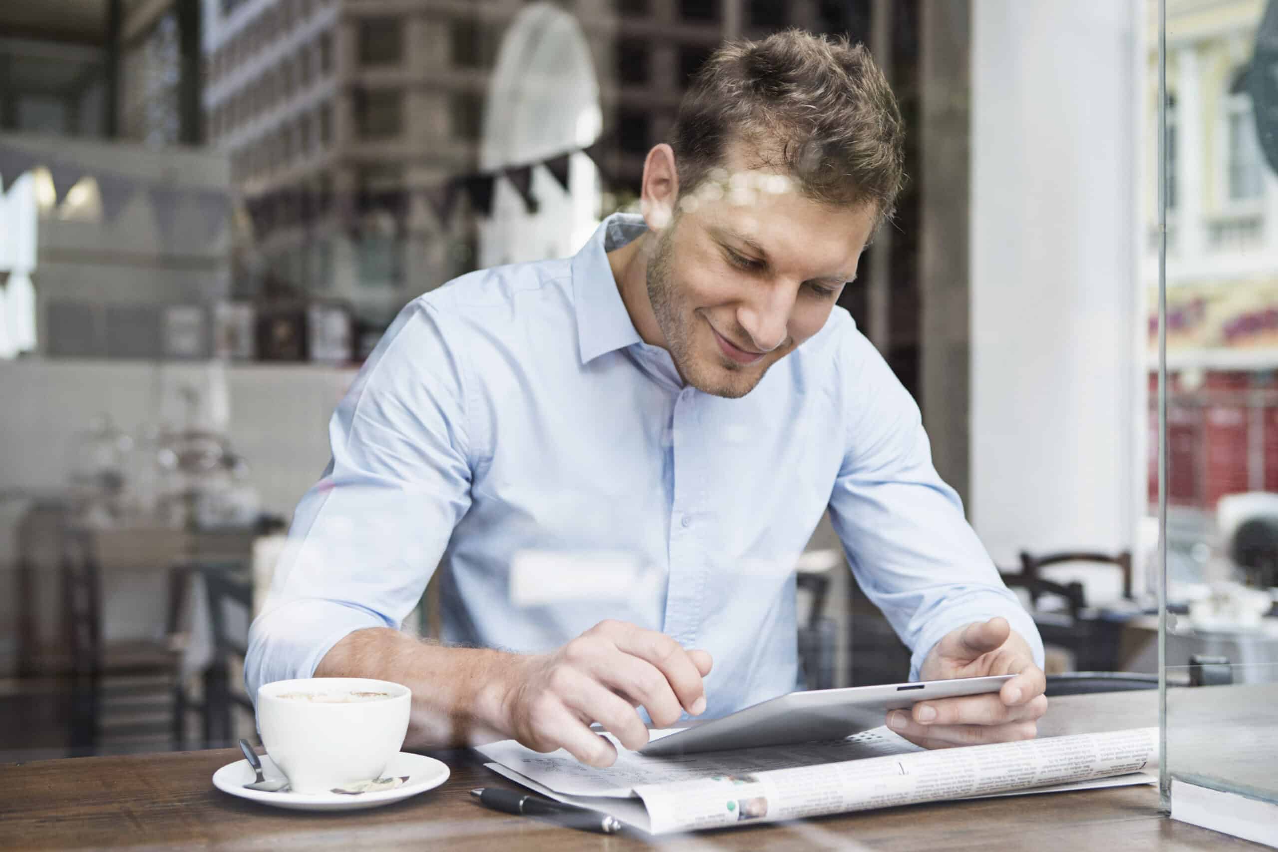 Ein Mann in einem hellblauen Hemd sitzt an einem Cafétisch und schaut auf ein Tablet. Neben ihm liegen eine Zeitung und eine Tasse Kaffee. In der hellen, modernen Umgebung wirkt er konzentriert und zufrieden. © Fotografie Tomas Rodriguez