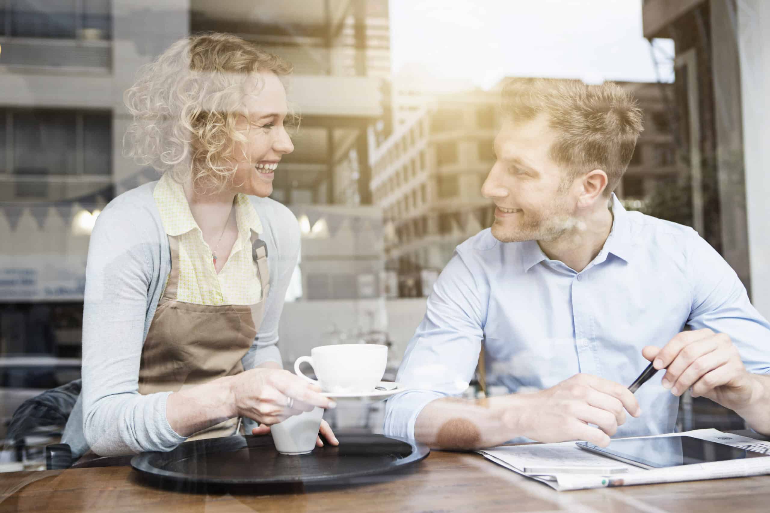Ein Mann und eine Frau unterhalten sich fröhlich bei einer Tasse Kaffee in einem Café. Die Frau trägt eine Barista-Schürze und der Mann ein Businesshemd. Sie sitzen an einem Tisch und legen eine Zeitung in die Mitte. © Fotografie Tomas Rodriguez