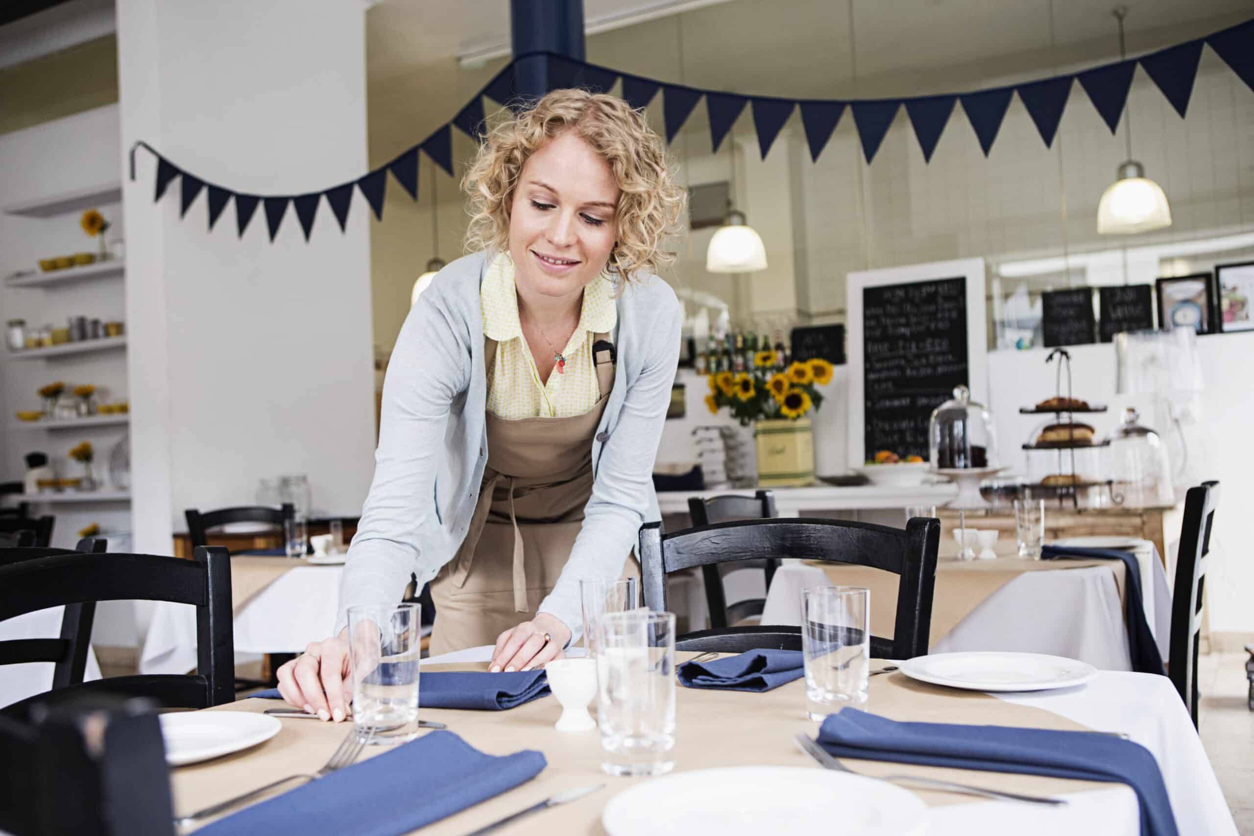 Eine Frau mit lockigem blondem Haar und einer Schürze deckt einen Tisch in einem hellen Café, das mit Sonnenblumen und Wimpel dekoriert ist. © Fotografie Tomas Rodriguez