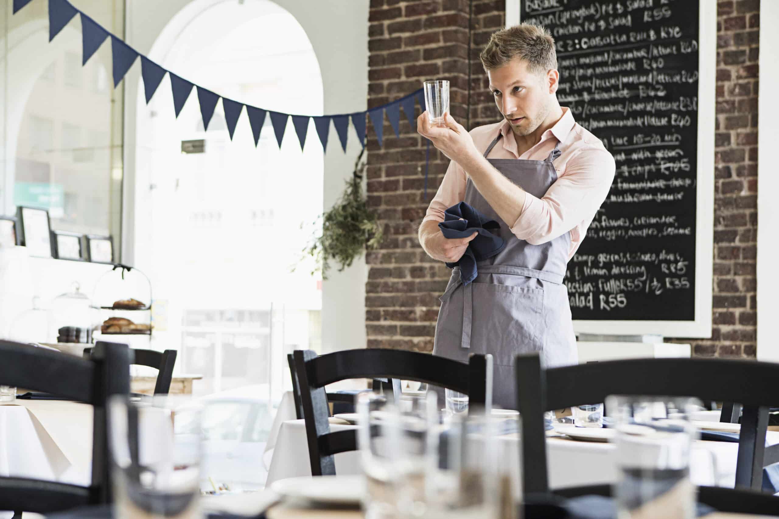 In einem gut beleuchteten Bistro überprüft ein Kellner die Sauberkeit eines Glases. Das mit Wimpel geschmückte Café bietet ordentlich angeordnete Tische und im Hintergrund eine Tafel mit der Speisekarte. © Fotografie Tomas Rodriguez