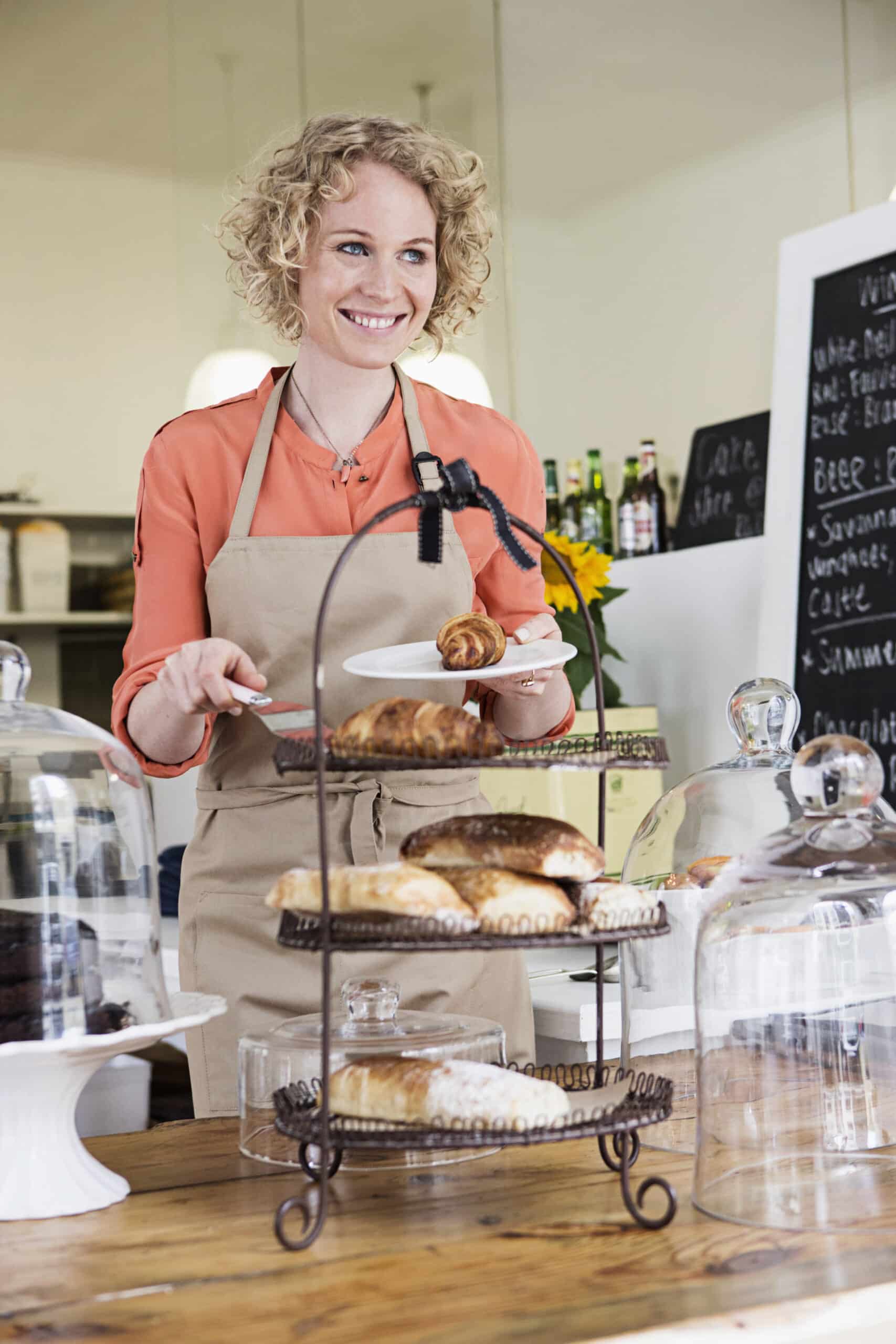Eine lächelnde Frau mit lockigem Haar und einer Schürze steht mit einer Zange und einem Croissant in einer Bäckerei. Vor ihr auf der Theke stehen Gläser mit Gebäck. © Fotografie Tomas Rodriguez