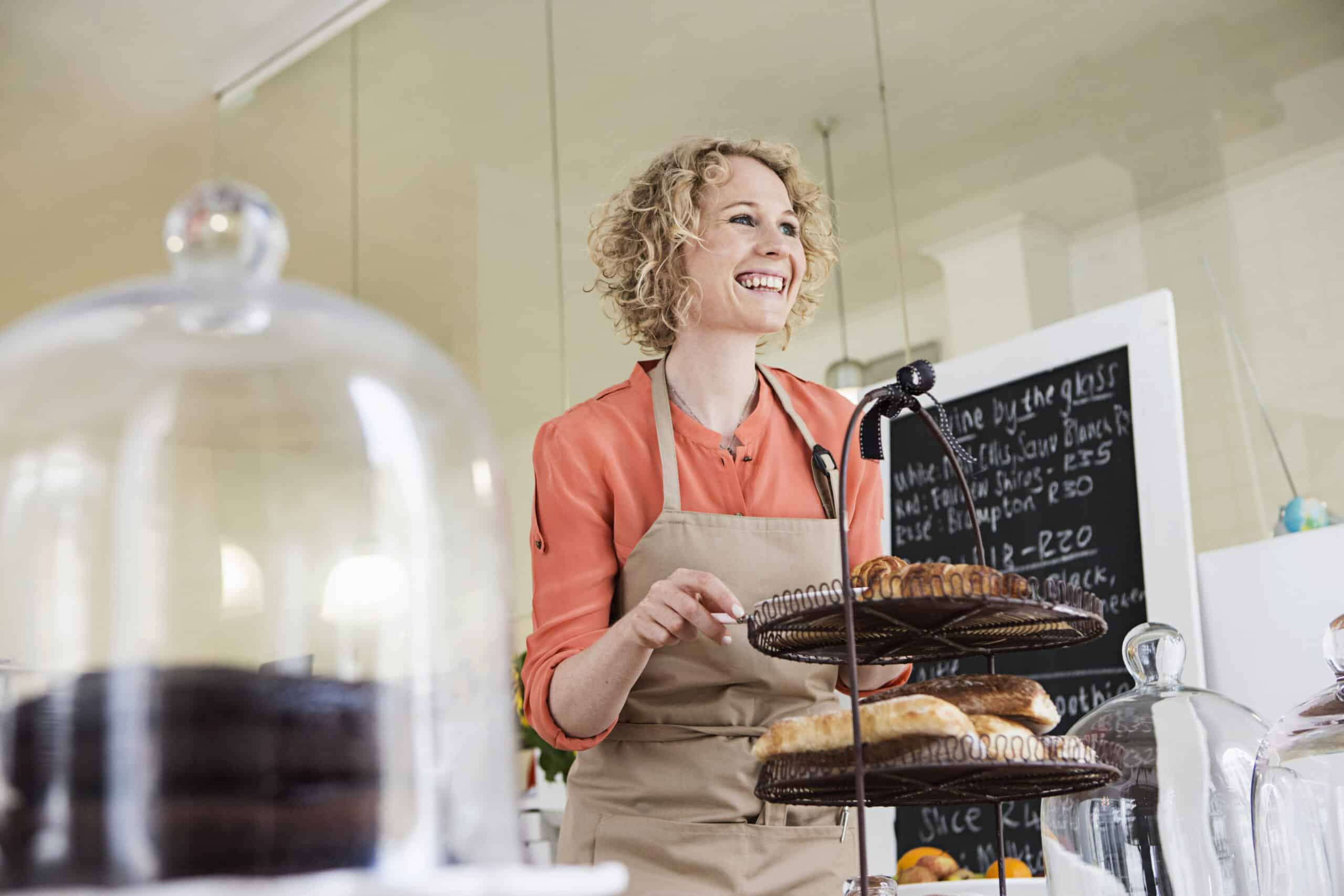 Eine fröhliche Frau in einer Schürze stellt ein Tablett mit frischem Gebäck auf die Theke eines Cafés, im Hintergrund ist eine Tafel mit der Speisekarte zu sehen. © Fotografie Tomas Rodriguez