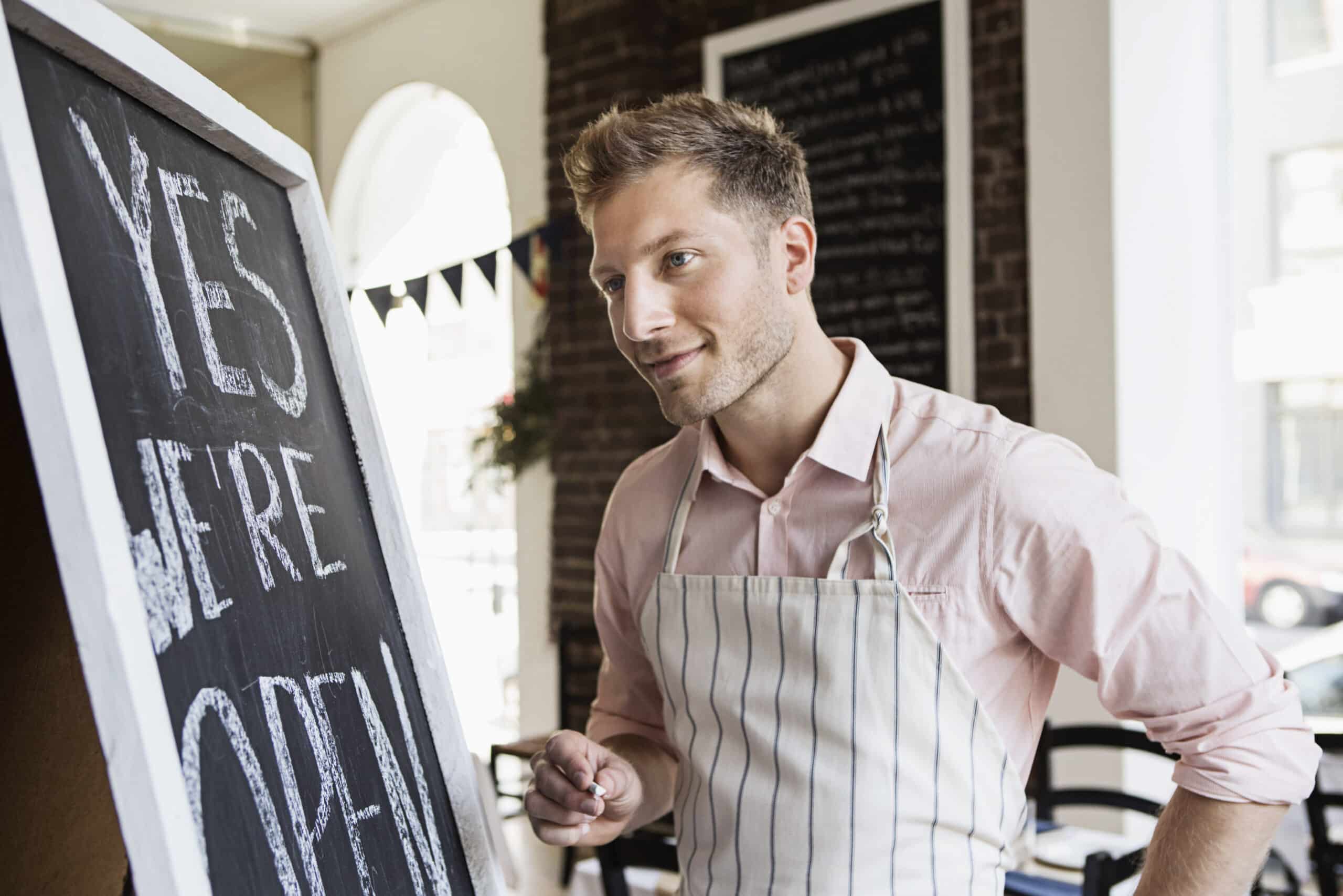 Ein lächelnder Cafébesitzer in einer gestreiften Schürze schreibt „Ja, wir haben geöffnet“ auf ein Kreidetafelschild in seinem hellen, einladenden Laden. © Fotografie Tomas Rodriguez