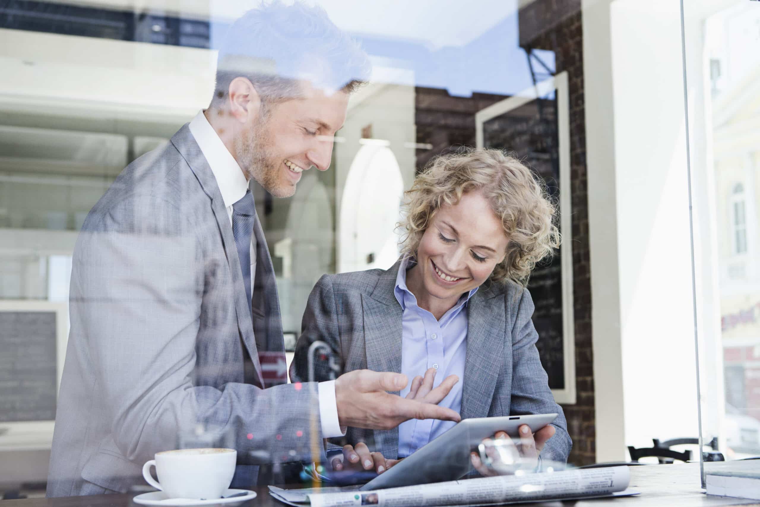 Zwei Geschäftsleute, ein Mann und eine Frau, lächeln und interagieren an einem Cafétisch über einem Tablet-PC, der Blick ist durch eine Glasscheibe möglich. © Fotografie Tomas Rodriguez