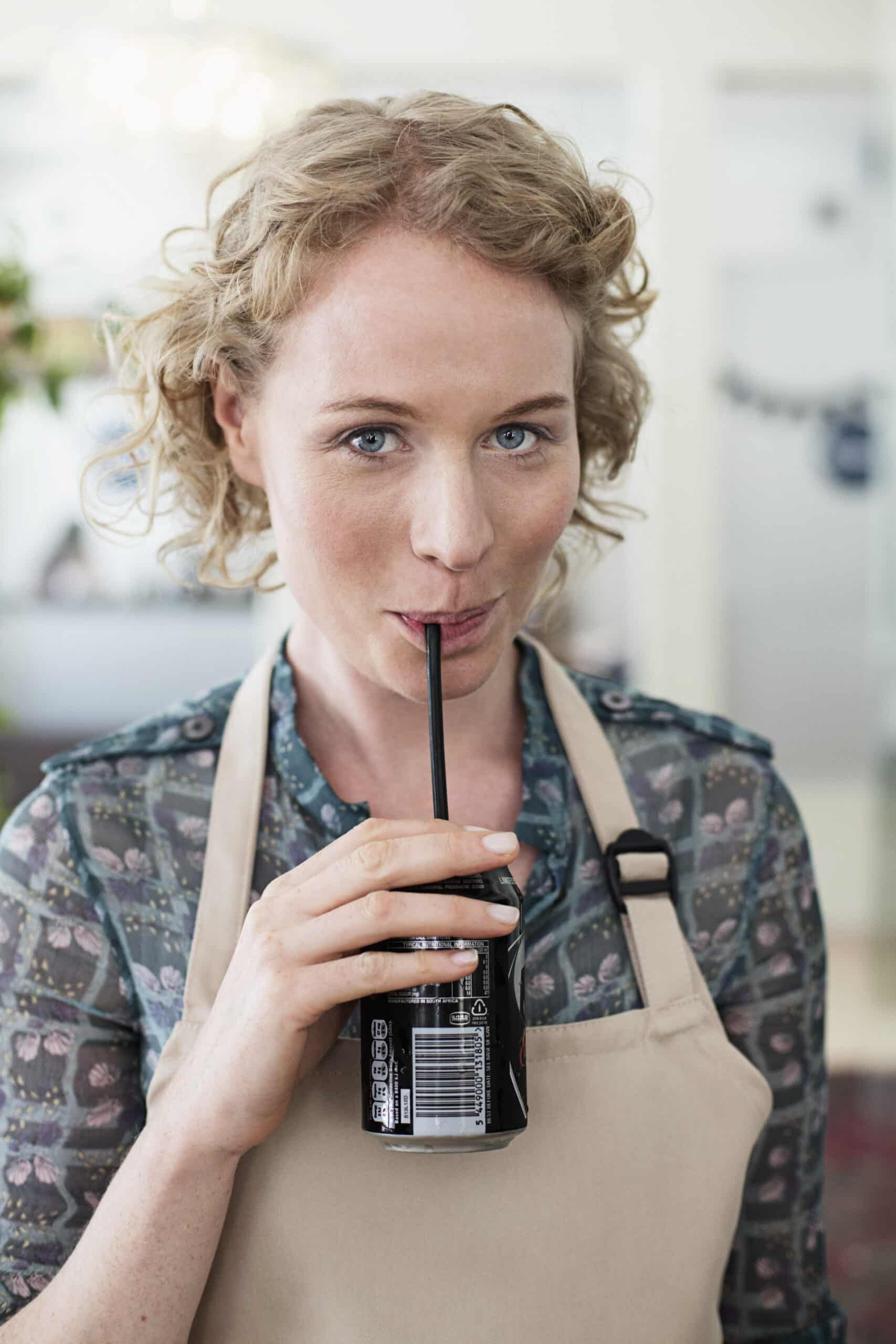 Eine Frau mit lockigem blondem Haar und einer geblümten Bluse, die eine Schürze trägt, nippt an einem Strohhalm aus einer Tasse und blickt mit einem subtilen Lächeln in die Kamera. © Fotografie Tomas Rodriguez