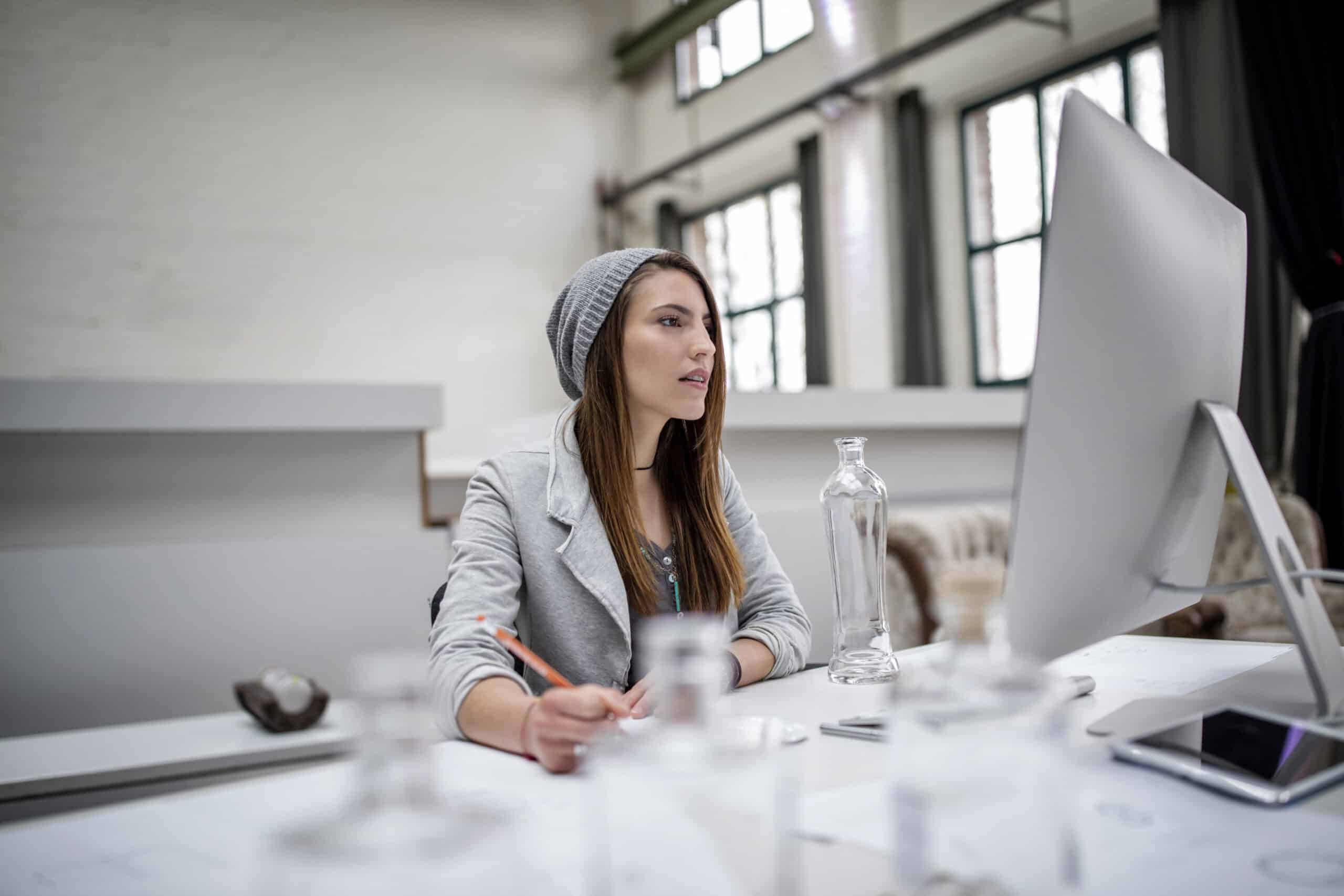 Eine junge Frau in einem grauen Blazer und einer Beanie-Mütze sitzt an einem Schreibtisch in einem hellen, modernen Büro und arbeitet konzentriert an einem Computer. Auf ihrem Schreibtisch liegen außerdem eine durchsichtige Wasserflasche und einige Unterlagen. © Fotografie Tomas Rodriguez