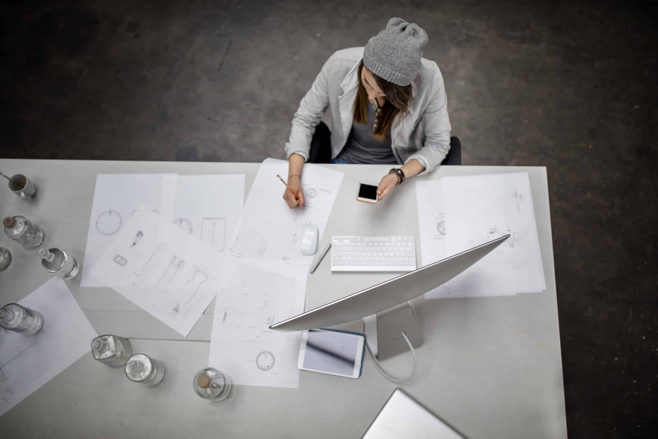 Eine Frau mit grauer Beanie-Mütze und weißem Hemd arbeitet an einem weißen Tisch, der mit Architekturzeichnungen, einem Smartphone und einem Laptop bedeckt ist (von oben gesehen). © Fotografie Tomas Rodriguez