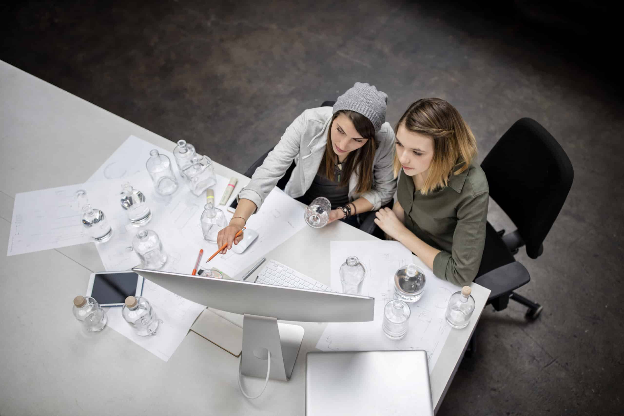 Zwei Frauen arbeiten gemeinsam an einem Schreibtisch voller Papiere und Glasflaschen an einem Computer in einer modernen Büroumgebung. © Fotografie Tomas Rodriguez