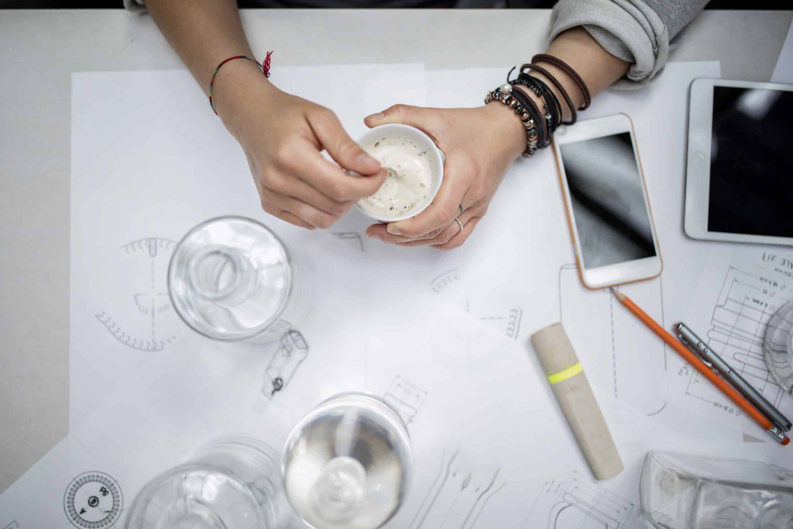 Draufsicht auf die Hände einer Person, die Materialien in einer Tasse mischt, umgeben von wissenschaftlichen Diagrammen, Gläsern, Bechern, einem Smartphone und einem Tablet auf einem weißen Tisch. © Fotografie Tomas Rodriguez