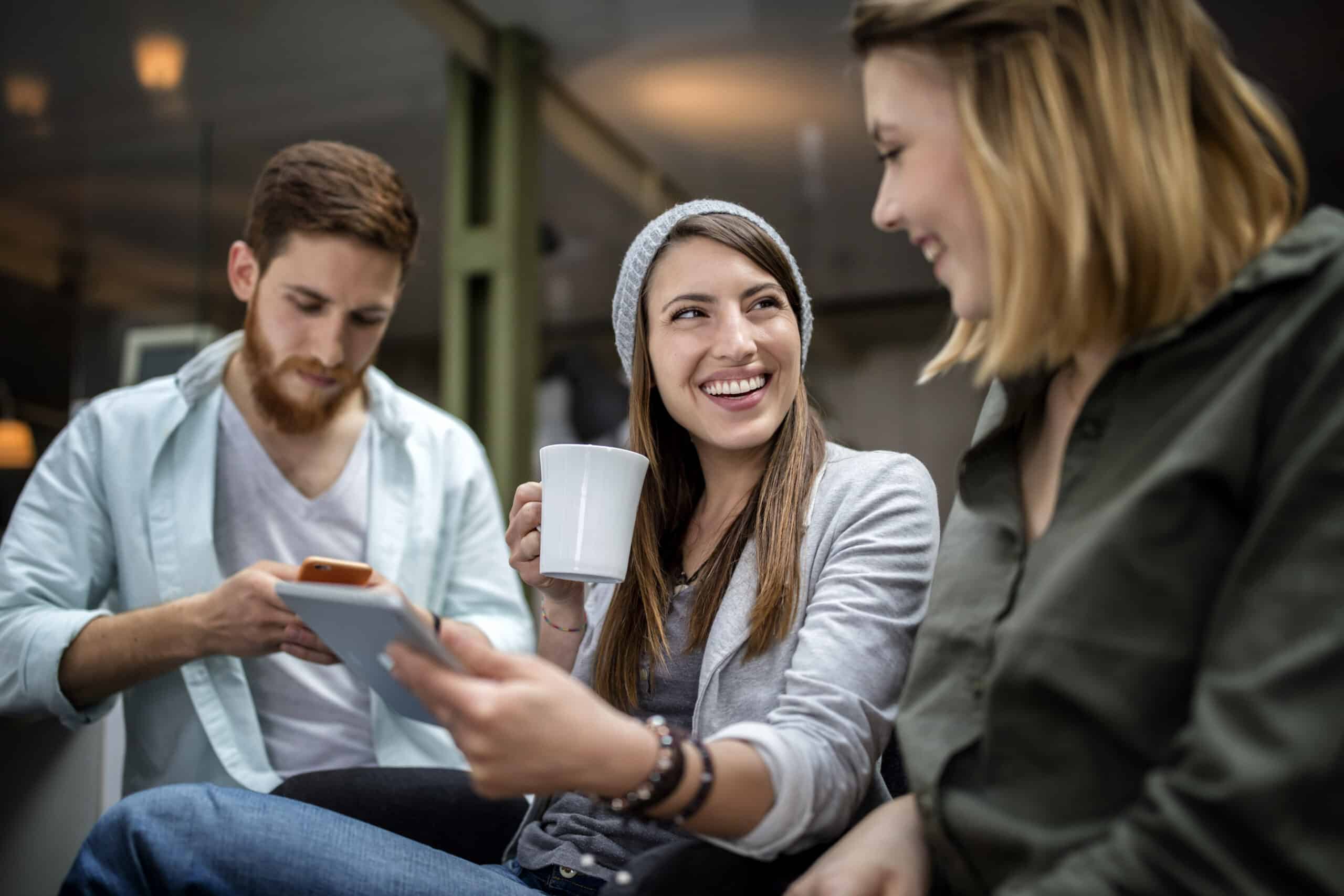 Drei Freunde genießen die gemeinsame Zeit in gemütlicher Atmosphäre; ein Mann ist auf sein Smartphone konzentriert, während zwei Frauen, eine davon mit einer Kaffeetasse in der Hand, lachen und angeregt plaudern. © Fotografie Tomas Rodriguez