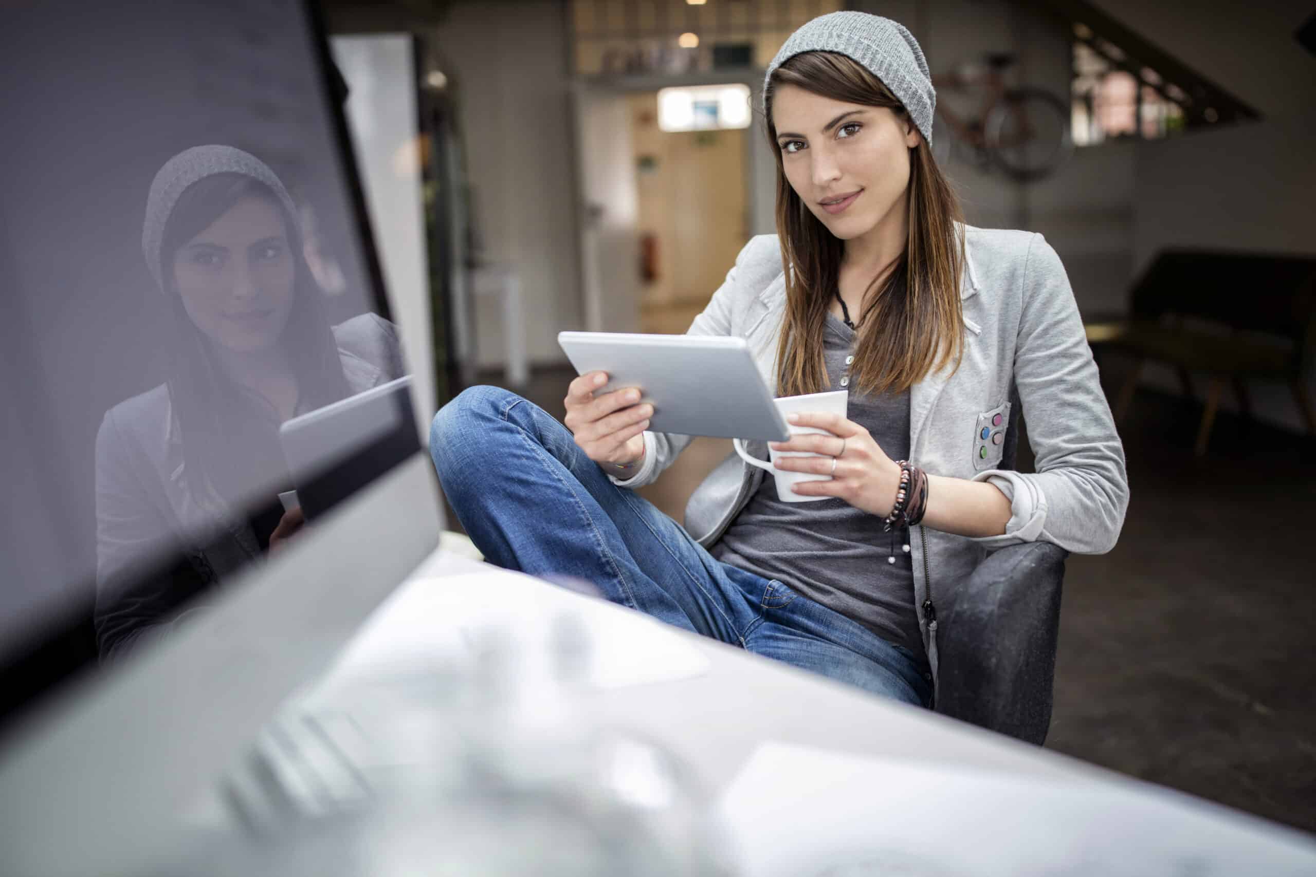 Eine junge Frau in einer lässigen grauen Jacke und Beanie-Mütze sitzt an einem Schreibtisch und benutzt ein Tablet mit einem reflektierenden Fenster, das im Hintergrund ihr Bild zeigt. Ein unscharfer Vordergrund suggeriert ein helles, modernes Interieur. © Fotografie Tomas Rodriguez