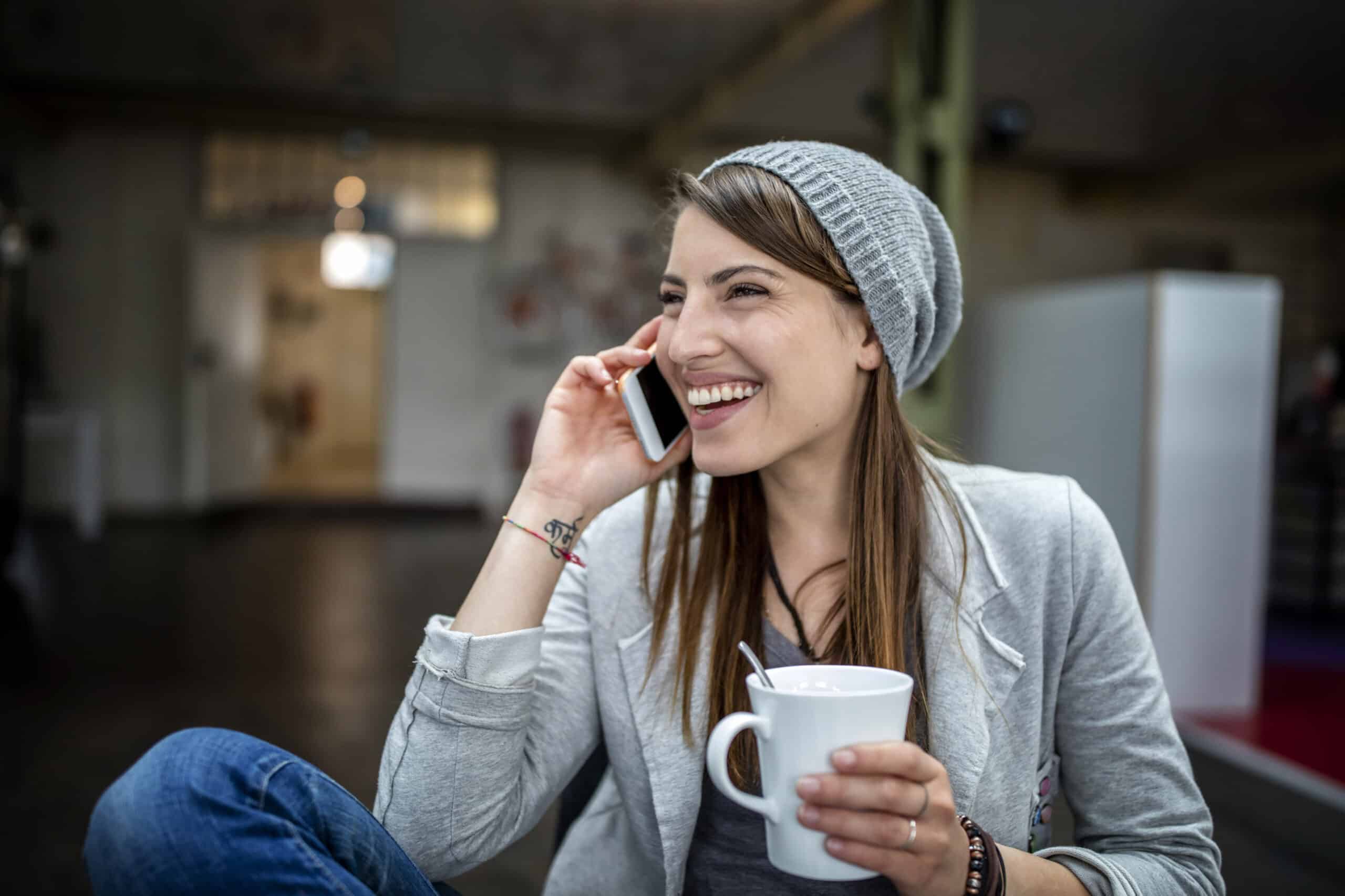Eine fröhliche junge Frau mit Beanie-Mütze telefoniert, während sie eine weiße Kaffeetasse hält und in einer ungezwungenen Büroumgebung sitzt. Sie lacht herzlich. © Fotografie Tomas Rodriguez