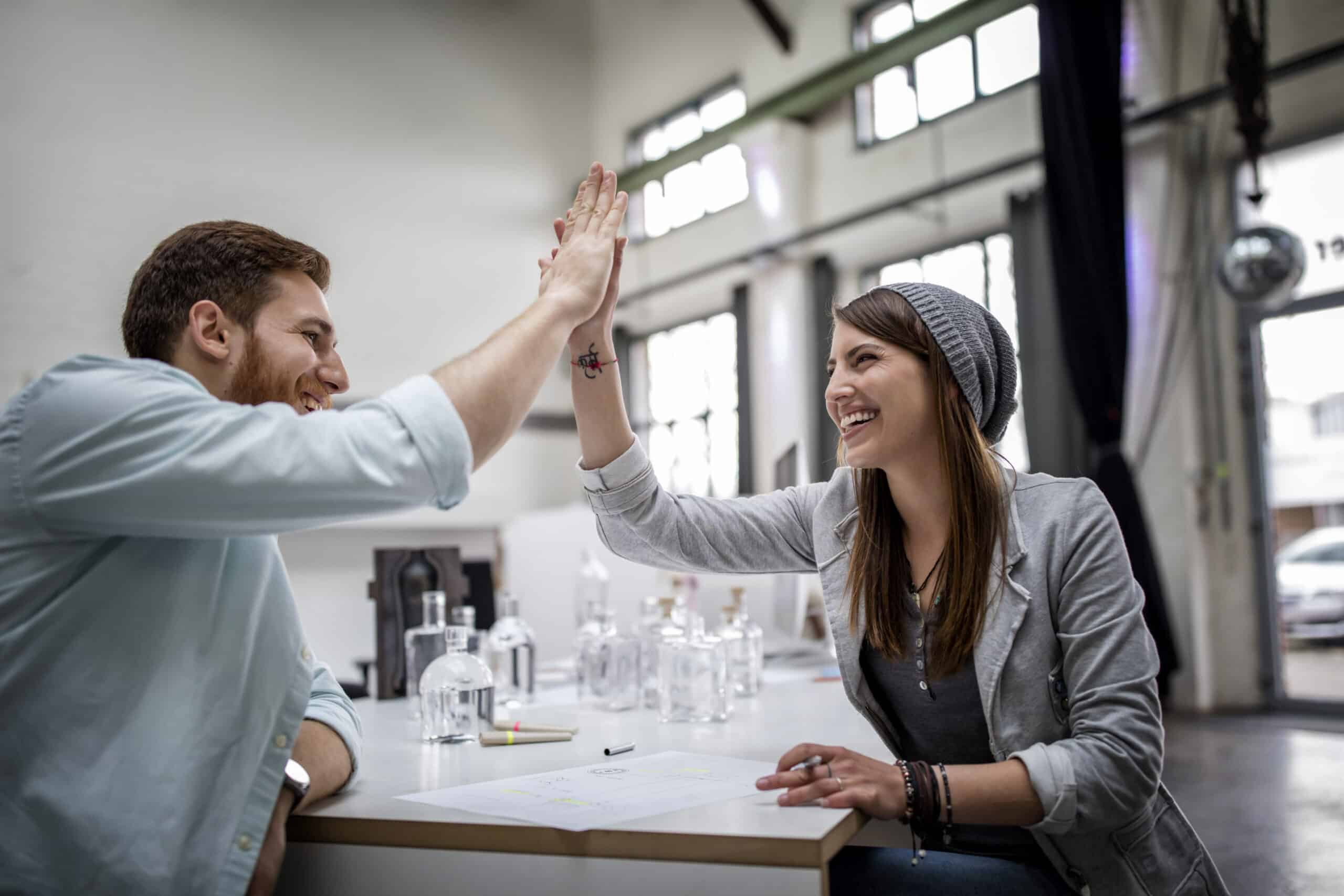 Zwei Kollegen, ein Mann und eine Frau, lächeln und geben sich gegenseitig ein High-Five an einem gemütlichen Büroarbeitsplatz mit Papieren und Laptops auf dem Tisch. © Fotografie Tomas Rodriguez