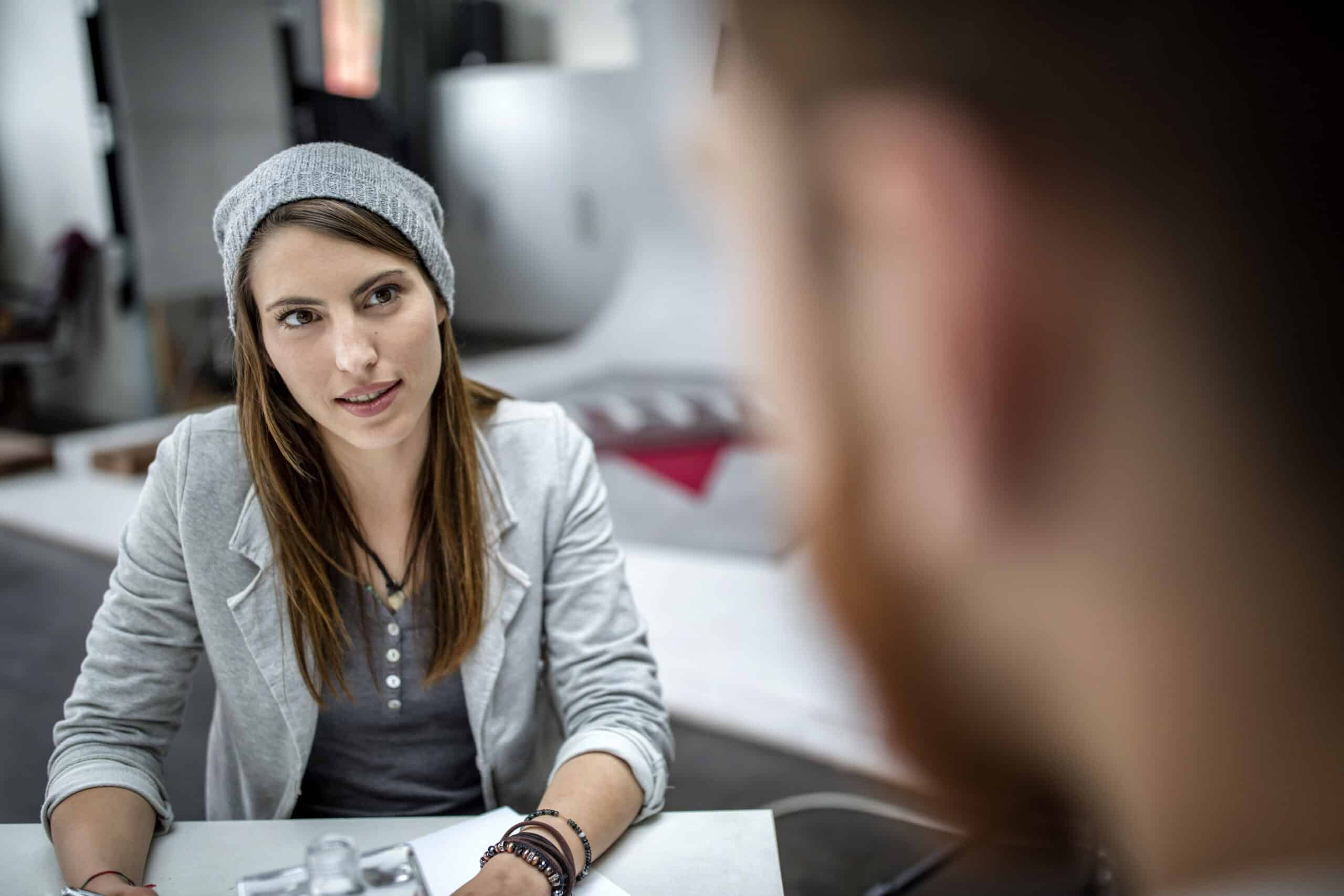 Eine junge Frau mit grauer Beanie-Mütze und hellgrauer Strickjacke sitzt an einem Tisch in einem Büro und unterhält sich mit einem ihr gegenüberstehenden Mann, dessen Bild nicht scharf abgebildet ist. © Fotografie Tomas Rodriguez