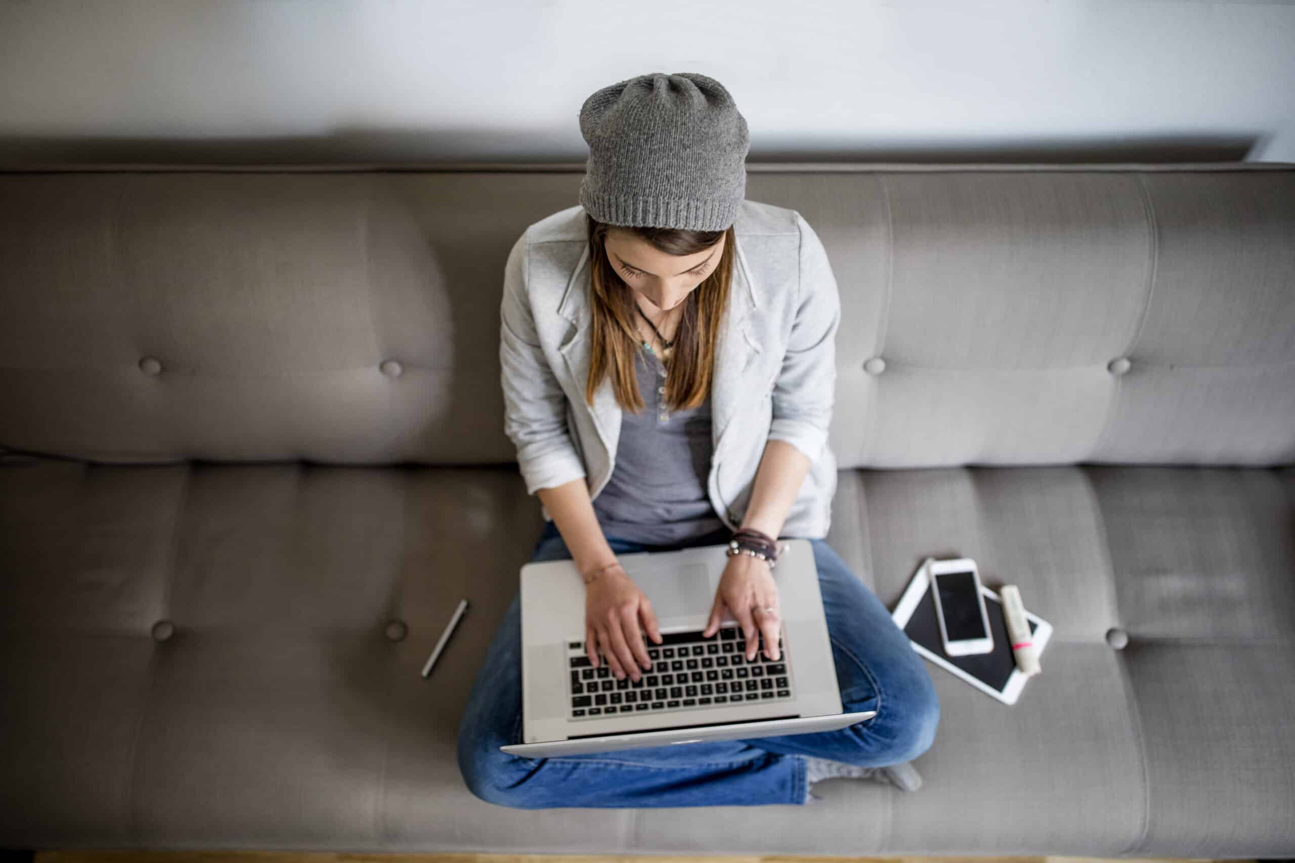 Eine junge Frau mit grauer Beaniemütze und weißem Pullover sitzt im Schneidersitz auf einem grauen Sofa und tippt auf einem Laptop, neben ihr liegen ein Smartphone und ein Tablet. © Fotografie Tomas Rodriguez