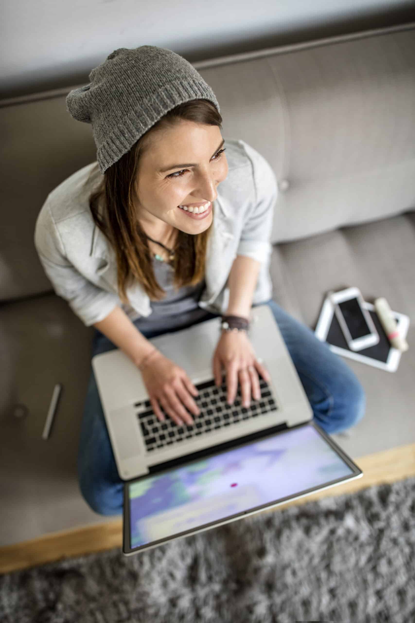 Eine junge Frau sitzt auf einem grauen Teppich und lächelt, während sie einen Laptop benutzt. Sie trägt eine hellgraue Mütze und Jacke. Neben ihr liegen ein Smartphone, ein Tablet und eine Smartwatch. © Fotografie Tomas Rodriguez