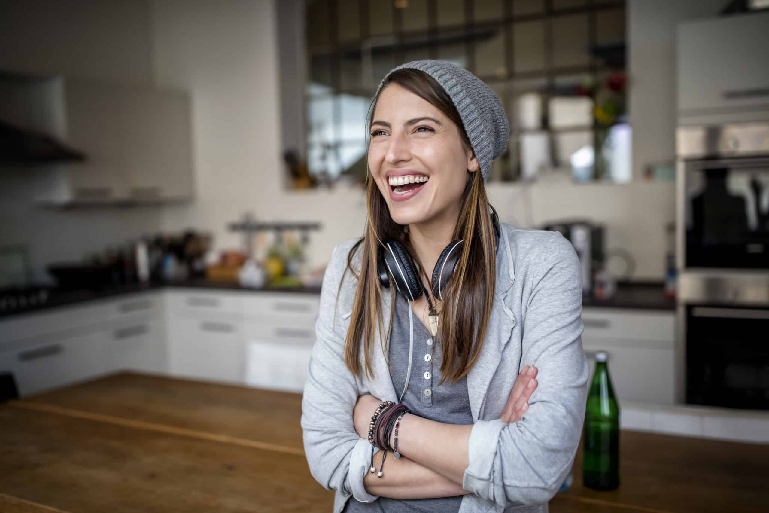Eine fröhliche Frau mit Beanie-Mütze und Kopfhörern um den Hals lacht, während sie mit verschränkten Armen in einer modernen Küche steht. © Fotografie Tomas Rodriguez