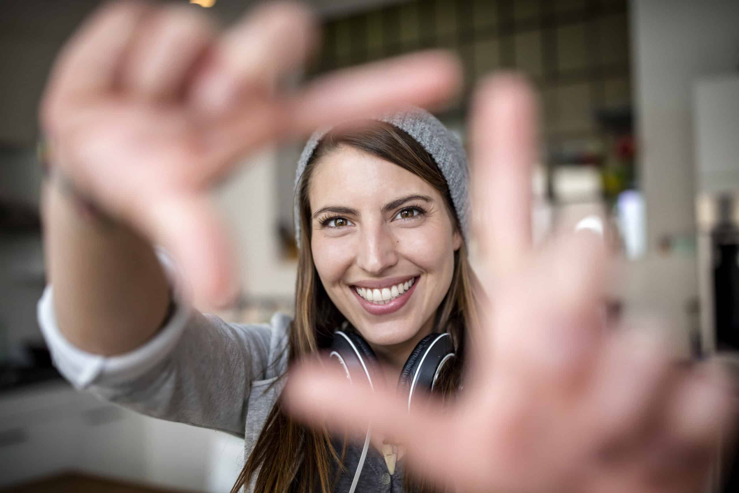 Eine fröhliche Frau mit Beanie und Kopfhörern rahmt mit ihren Fingern einen Kameraschuss ein und lächelt in einer hellen, modernen Küche. © Fotografie Tomas Rodriguez