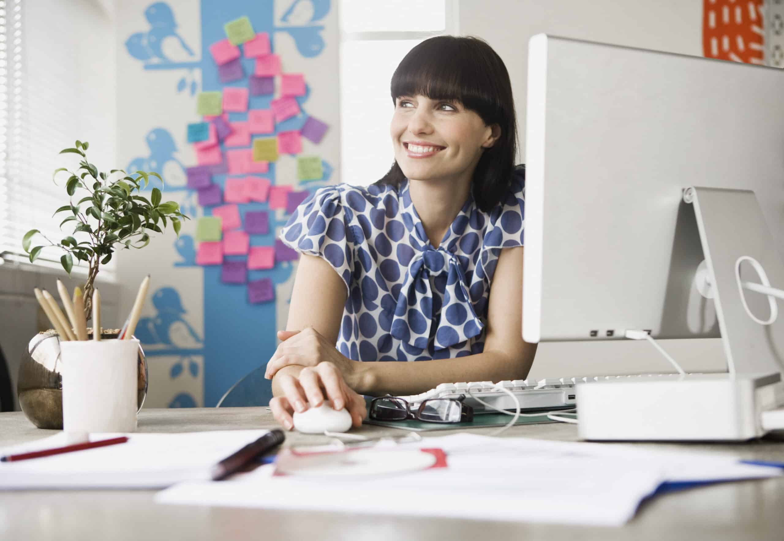 Eine lächelnde Frau in einer blauen Bluse mit Tupfen sitzt mit einer Computermaus in der Hand an einem Schreibtisch. Auf dem Schreibtisch stehen ein Computermonitor, eine Pflanze und verschiedene Büromaterialien. Im Hintergrund ist eine bunte Wand mit Haftnotizen zu sehen. © Fotografie Tomas Rodriguez