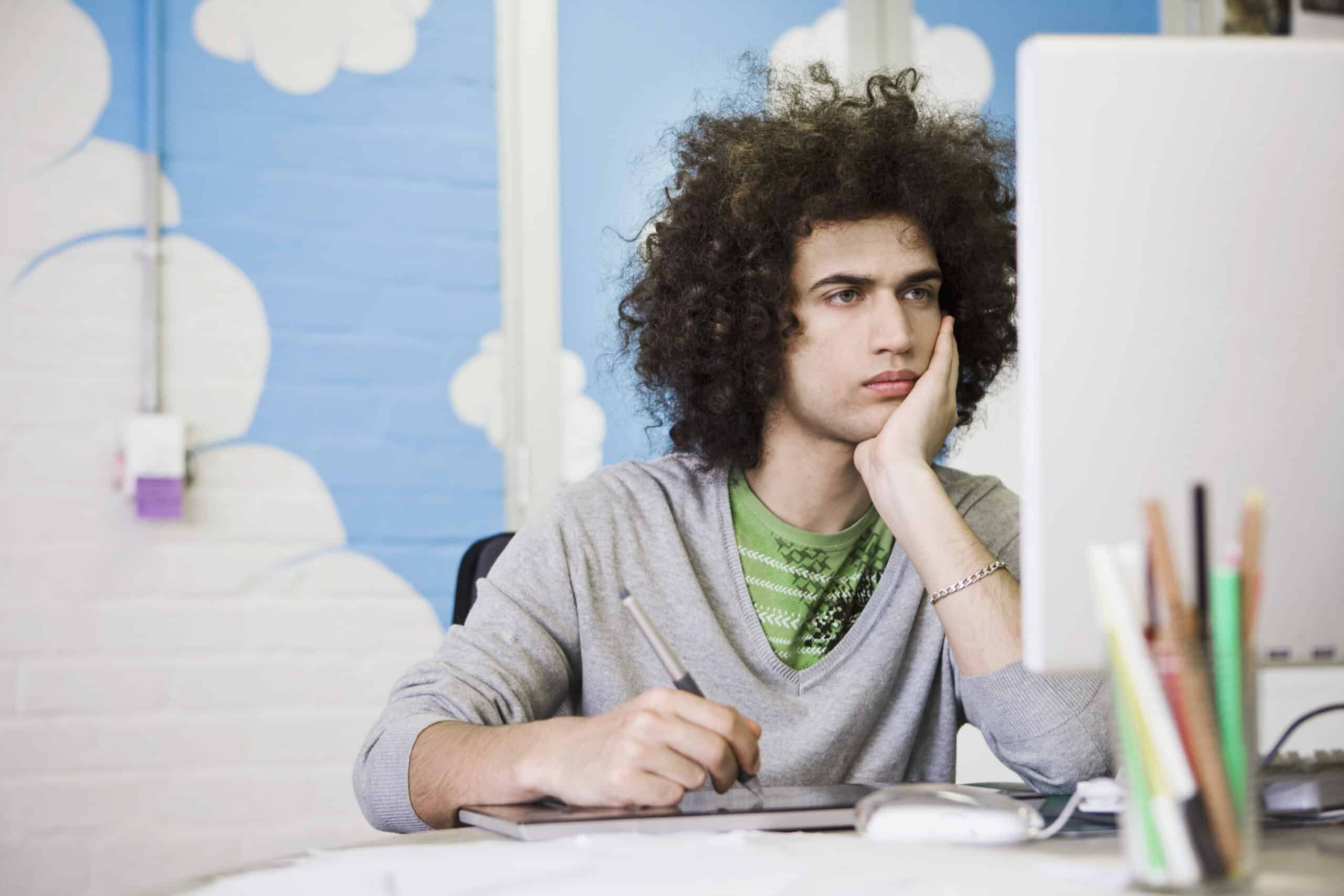 Eine Person mit lockigem Haar sitzt an einem Schreibtisch und starrt auf einen Computerbildschirm, während sie ihren Kopf auf eine Hand stützt. In der anderen Hand hält sie einen Stift. Im Hintergrund ist eine blaue Wand mit weißen Wolkendekorationen zu sehen. Auf dem Schreibtisch liegen einige Papiere und Schreibwaren. © Fotografie Tomas Rodriguez
