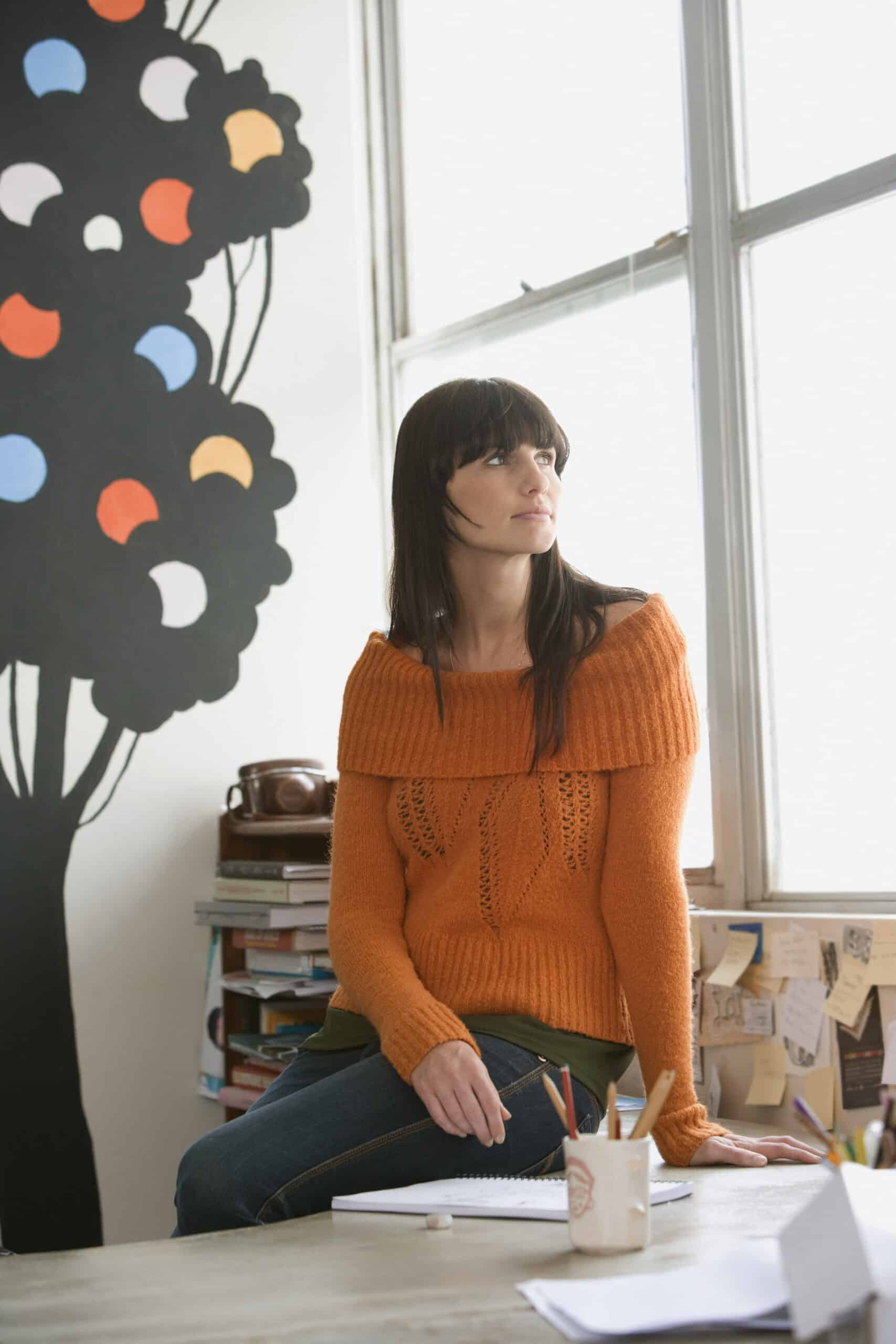 Eine Frau mit langen dunklen Haaren und einem orangefarbenen Pullover sitzt mit einem Stift in der Hand auf der Kante eines Schreibtischs. Sie blickt aus dem Fenster, hinter ihr an der Wand ein gemaltes Baumbild mit bunten Punkten. An ihrem Arbeitsplatz liegen Bücher und Haftnotizen. © Fotografie Tomas Rodriguez
