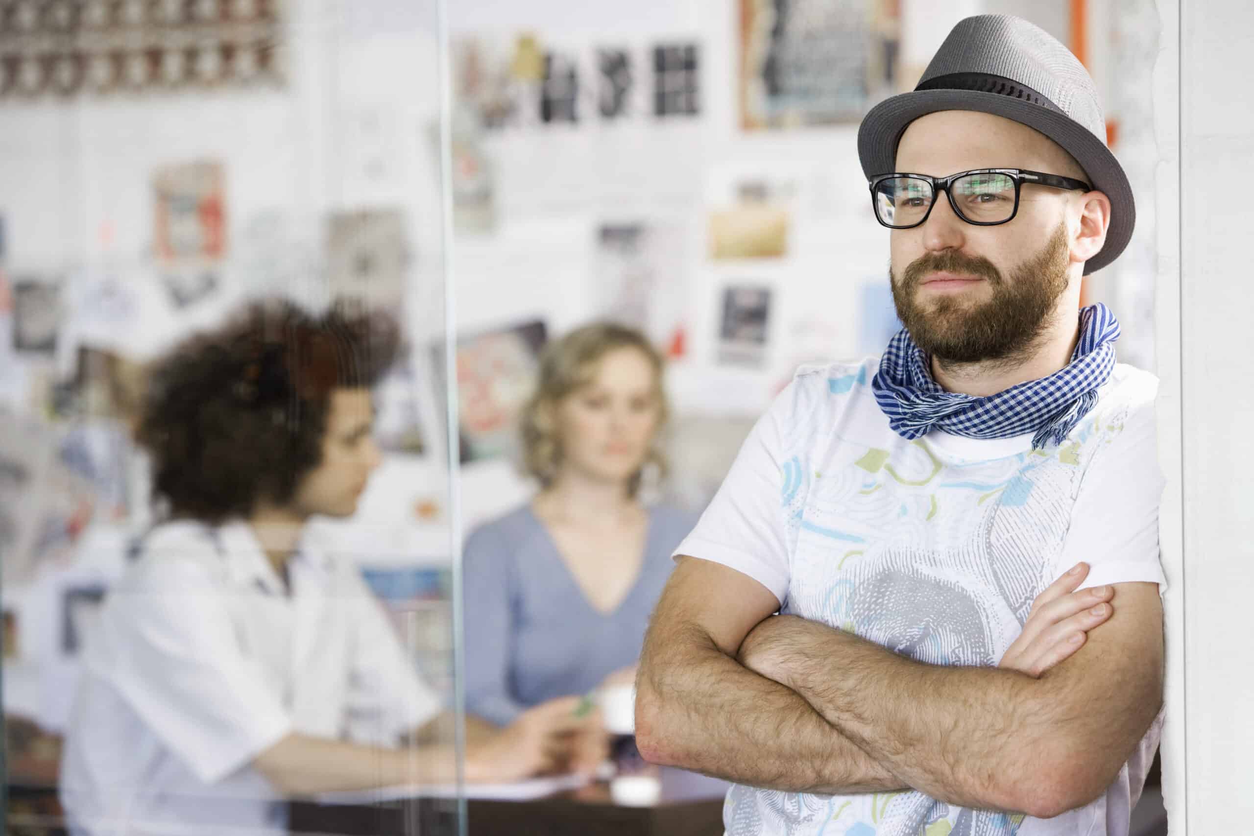 Ein bärtiger Mann mit Brille, Mütze und Schal steht mit verschränkten Armen an einer Glaswand gelehnt. Im unscharfen Hintergrund sitzen zwei Personen, eine mit lockigem Haar und eine mit blondem Haar, an einem Tisch in einem kreativen Büroraum. © Fotografie Tomas Rodriguez