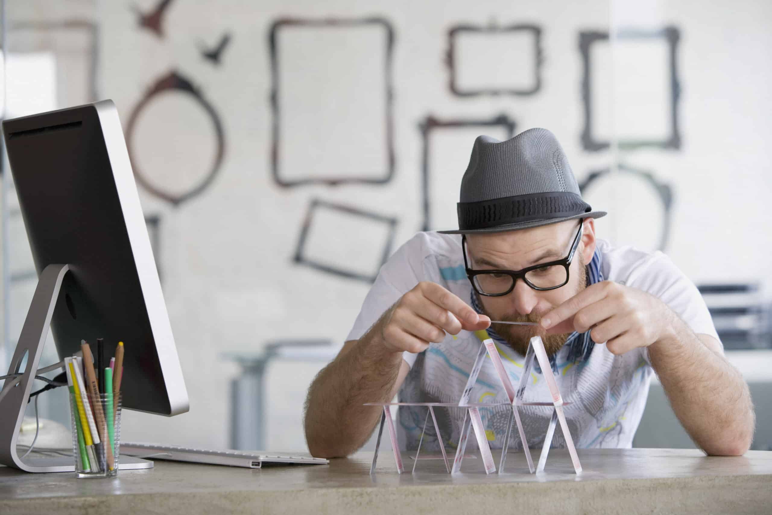 Ein Mann mit Brille und grauem Hut baut sorgfältig ein Kartenhaus auf einem Schreibtisch. Auf dem Schreibtisch liegen außerdem Bleistifte in einem Halter und ein Computermonitor. An der Wand hinter ihm sind mehrere leere Bilderrahmen zu sehen. © Fotografie Tomas Rodriguez