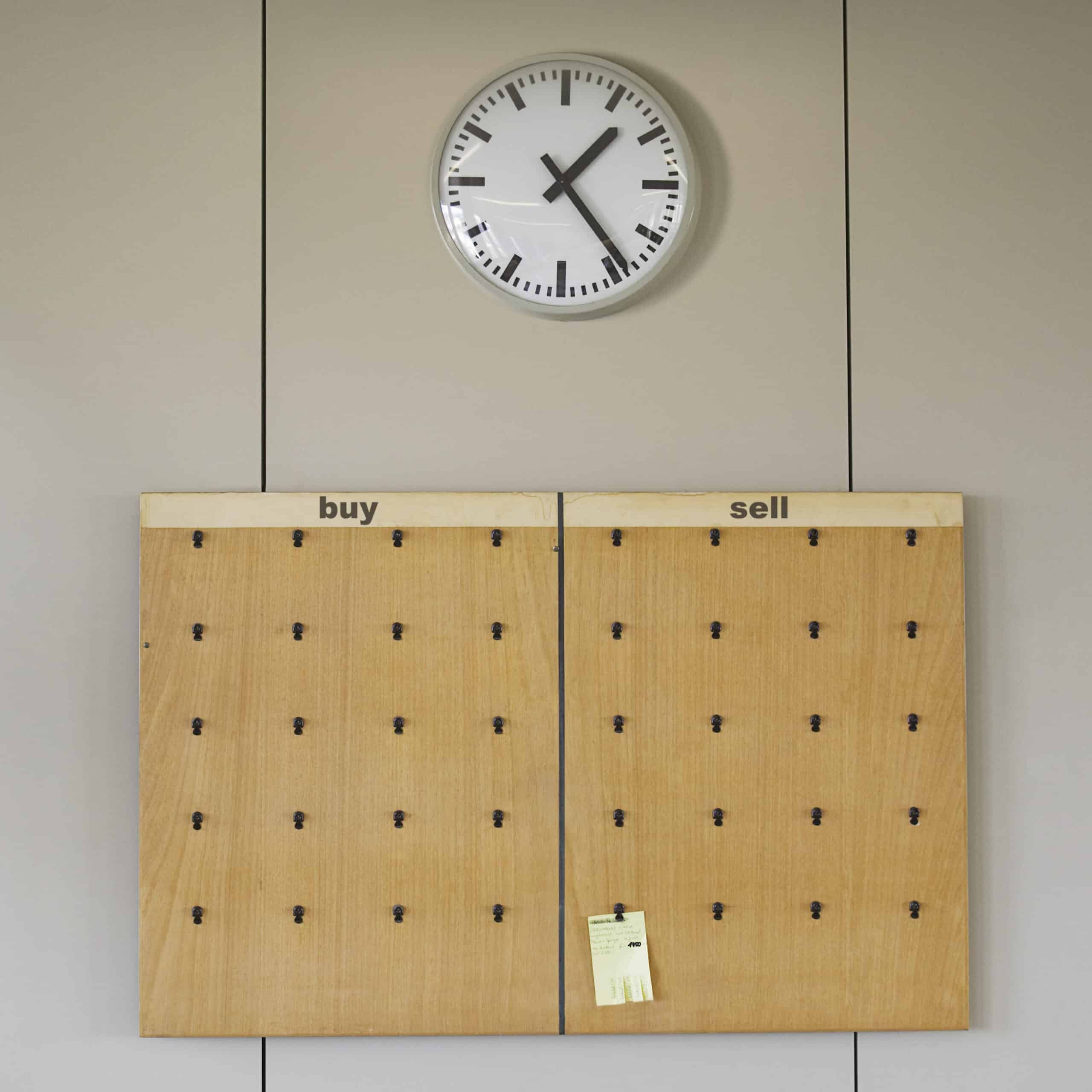 Ein Brett mit den Rubriken „Kaufen“ und „Verkaufen“ enthält Haken für Aushänge, aber nur ein Zettel ist befestigt. Über dem Brett hängt eine Uhr an der Wand, die 1:05 Uhr anzeigt. Der Hintergrund besteht aus schlichten, hellen Tafeln. © Fotografie Tomas Rodriguez