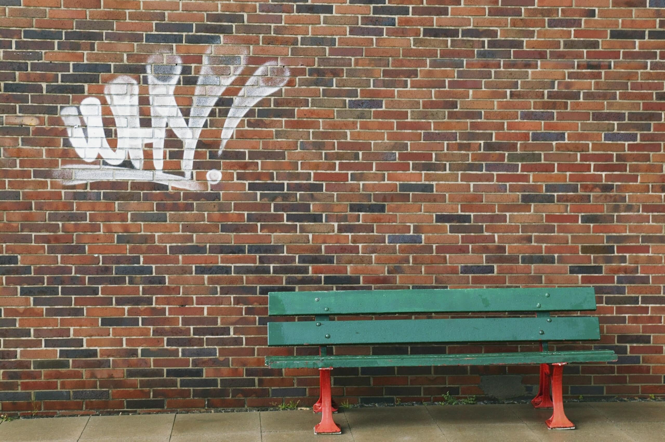 Eine grüne Holzbank mit roten Beinen steht vor einer Backsteinmauer mit Graffiti. Das Graffiti in weißer Farbe ergibt den Ausdruck „WAY!“. Der Boden davor ist gepflastert und leicht feucht, was auf kürzlichen Regen schließen lässt. © Fotografie Tomas Rodriguez