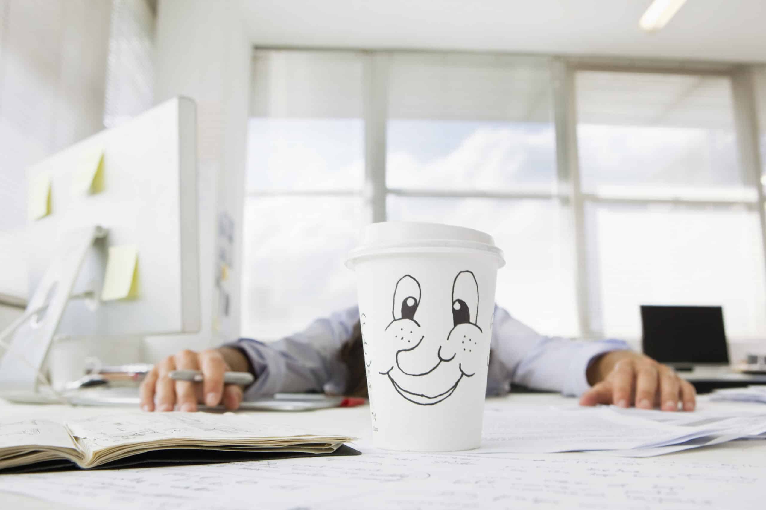 Eine Kaffeetasse mit einem lächelnden Cartoon-Gesicht steht auf einem unordentlichen Schreibtisch, auf dem offene Notizbücher und verstreute Papiere liegen. Im Hintergrund sitzt eine Person mit ausgestreckten Armen am Schreibtisch, teilweise verdeckt durch die Tasse, in einem hell erleuchteten Büro. © Fotografie Tomas Rodriguez