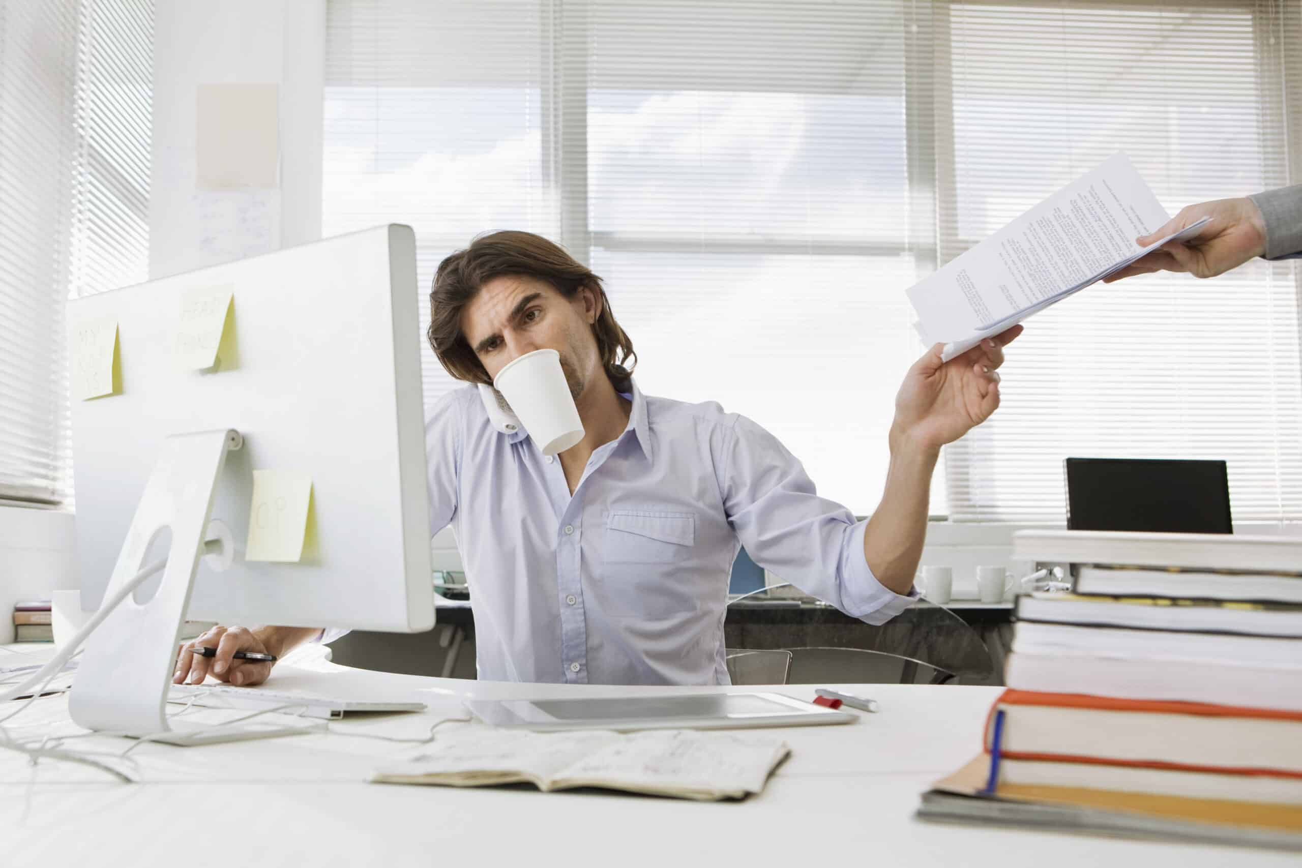 Eine Person am Schreibtisch erledigt mehrere Dinge gleichzeitig, schaut auf einen Computermonitor, hält in einer Hand ein Dokument und hat eine Kaffeetasse im Mund. Sie sitzt in einem hellen Büro mit Stapeln von Büchern und Papieren auf dem Schreibtisch, und eine andere Hand reicht ihr weitere Dokumente. © Fotografie Tomas Rodriguez