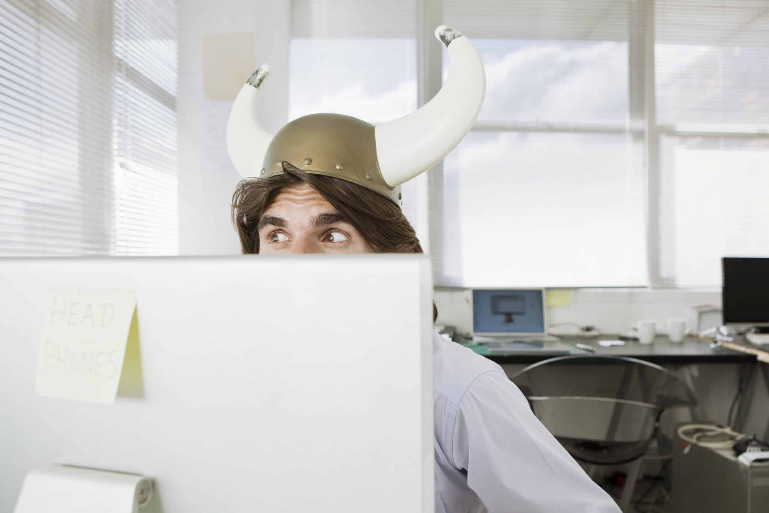 Eine Person mit einem Wikingerhelm mit weißen Hörnern blickt über einen Computermonitor in einem hell erleuchteten Büro. An der Rückseite des Monitors klebt ein Haftzettel mit der Aufschrift „KOPFHÖRER“. © Fotografie Tomas Rodriguez