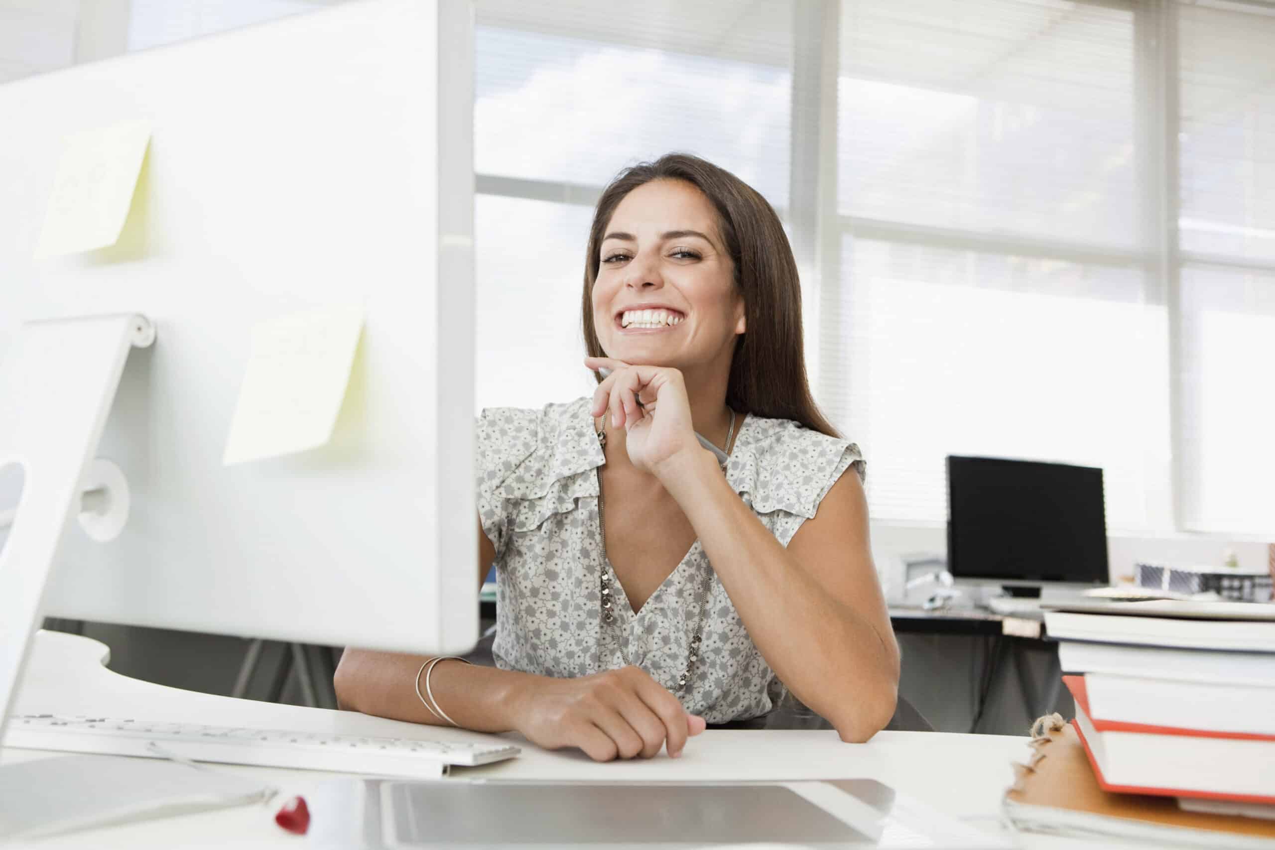 Eine Frau mit langen braunen Haaren und einer weiß gemusterten Bluse sitzt an einem Schreibtisch in einem Büro. Sie lächelt und schaut auf einen Computerbildschirm vor ihr. Auf dem Schreibtisch liegen verschiedene Büromaterialien und Akten, im Hintergrund sind unscharfe Bürogeräte zu sehen. © Fotografie Tomas Rodriguez